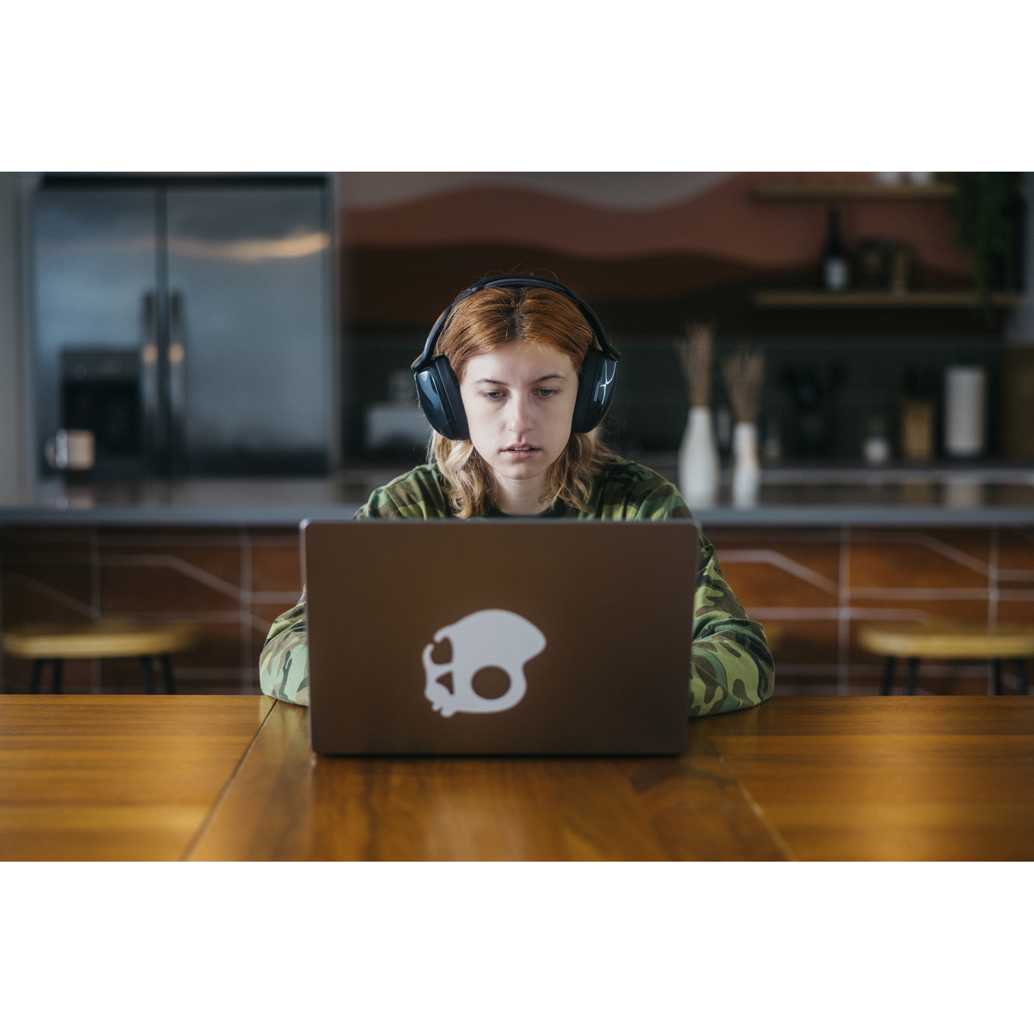 Une jeune femme avec un casque regarde un ordinateur portable. Elle porte une chemise de camouflage. Le fond est une cuisine.