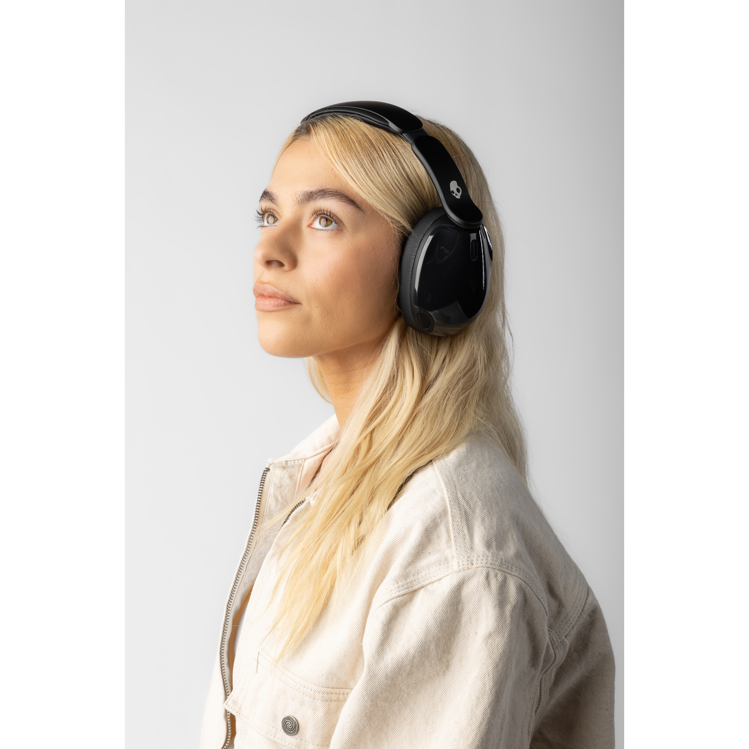 Femme blonde portant un casque noir et une veste crème. Elle regarde vers le haut sur fond blanc.