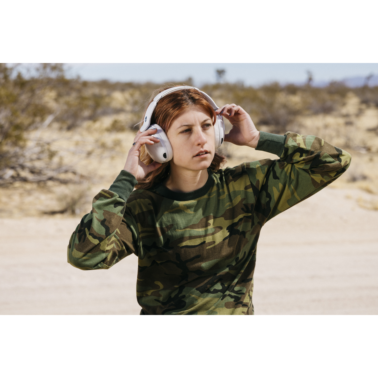 Une femme en chemise camouflage ajuste un casque blanc. Arrière-plan désertique avec buissons et ciel pâle.