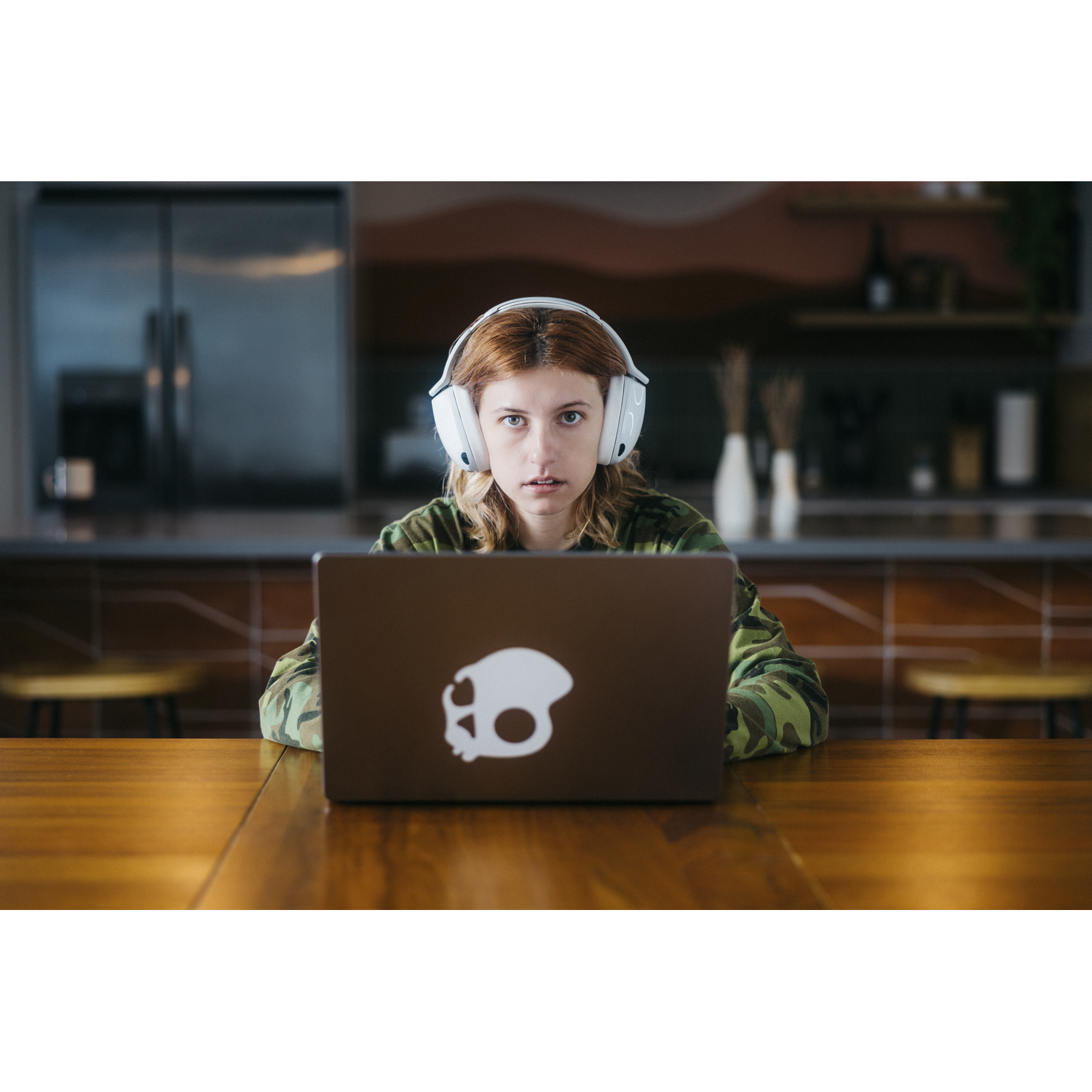 Une femme portant un casque regarde le spectateur par-dessus un ordinateur portable. L'ordinateur portable a un logo de crâne. Elle est assise à une table en bois.