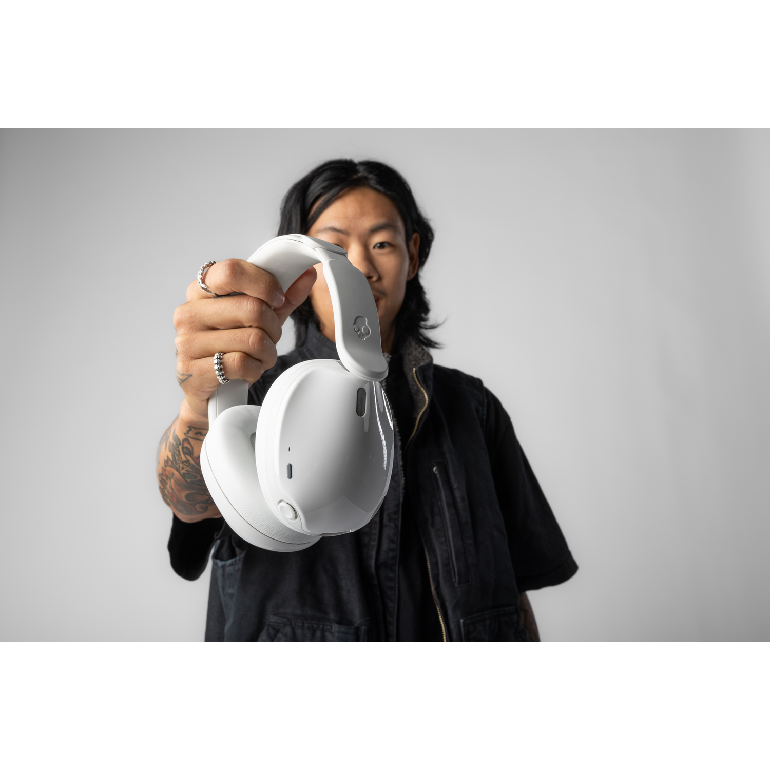Une personne tient un casque blanc. Il a des tatouages sur le bras et porte une veste noire. Le fond est gris uni.