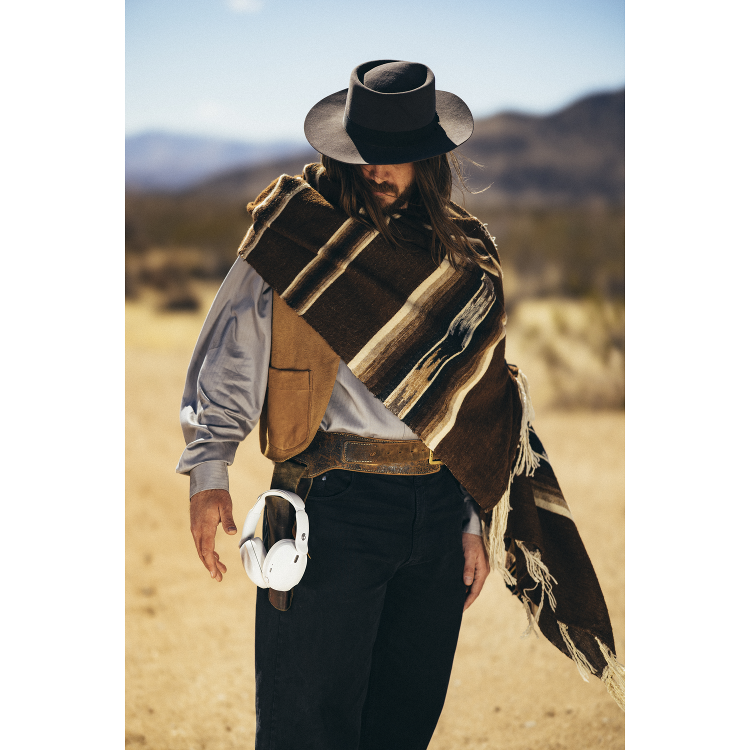 Un homme en chapeau de cowboy et poncho se tient dans le désert. Il porte un gilet, a les cheveux longs et tient un casque blanc.