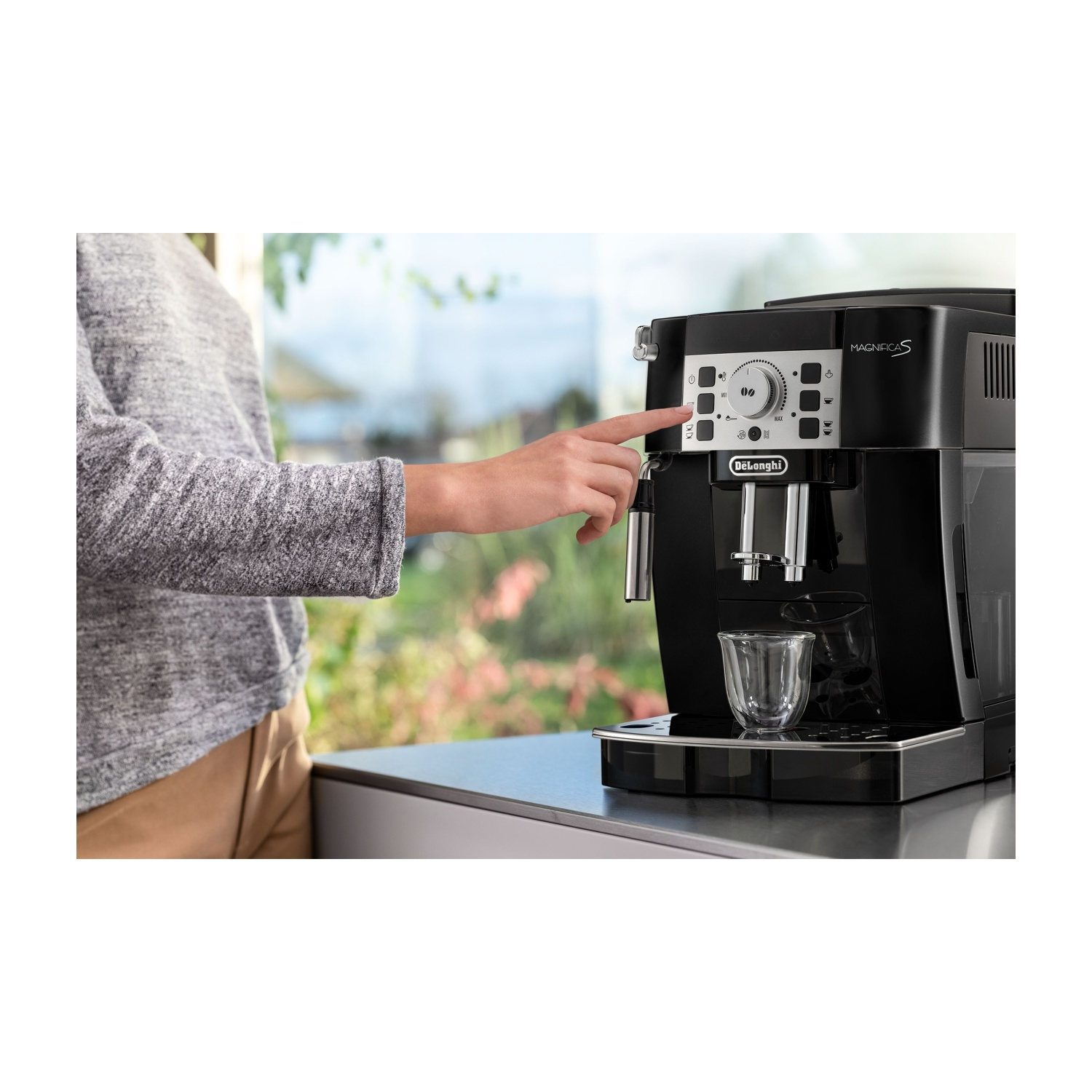 Una persona preme il pulsante su una macchina da caffè nera. Un bicchiere è sotto l'erogatore. Lo sfondo mostra verde attraverso una finestra.