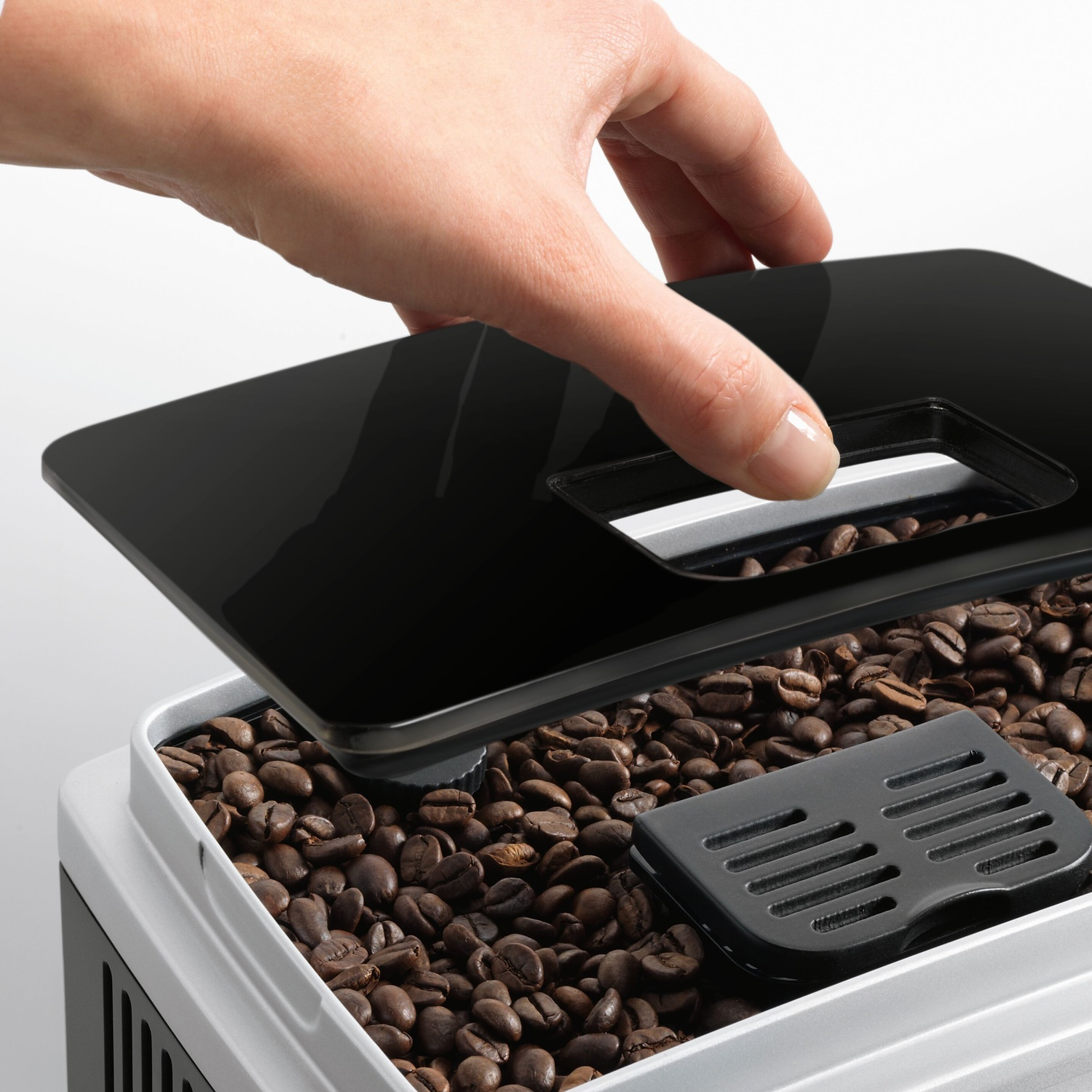 Una mano apre un coperchio nero rivelando chicchi di caffè. Un vassoio nero all'interno. Macchina bianca e nera con sfondo bianco.