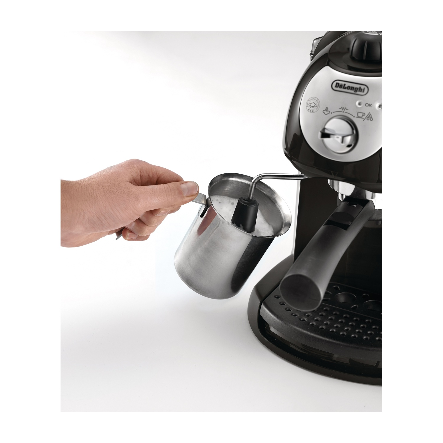 Una mano tiene un bricco in acciaio per montare il latte. A destra, una macchina per caffè espresso De'Longhi nera. Sfondo bianco.