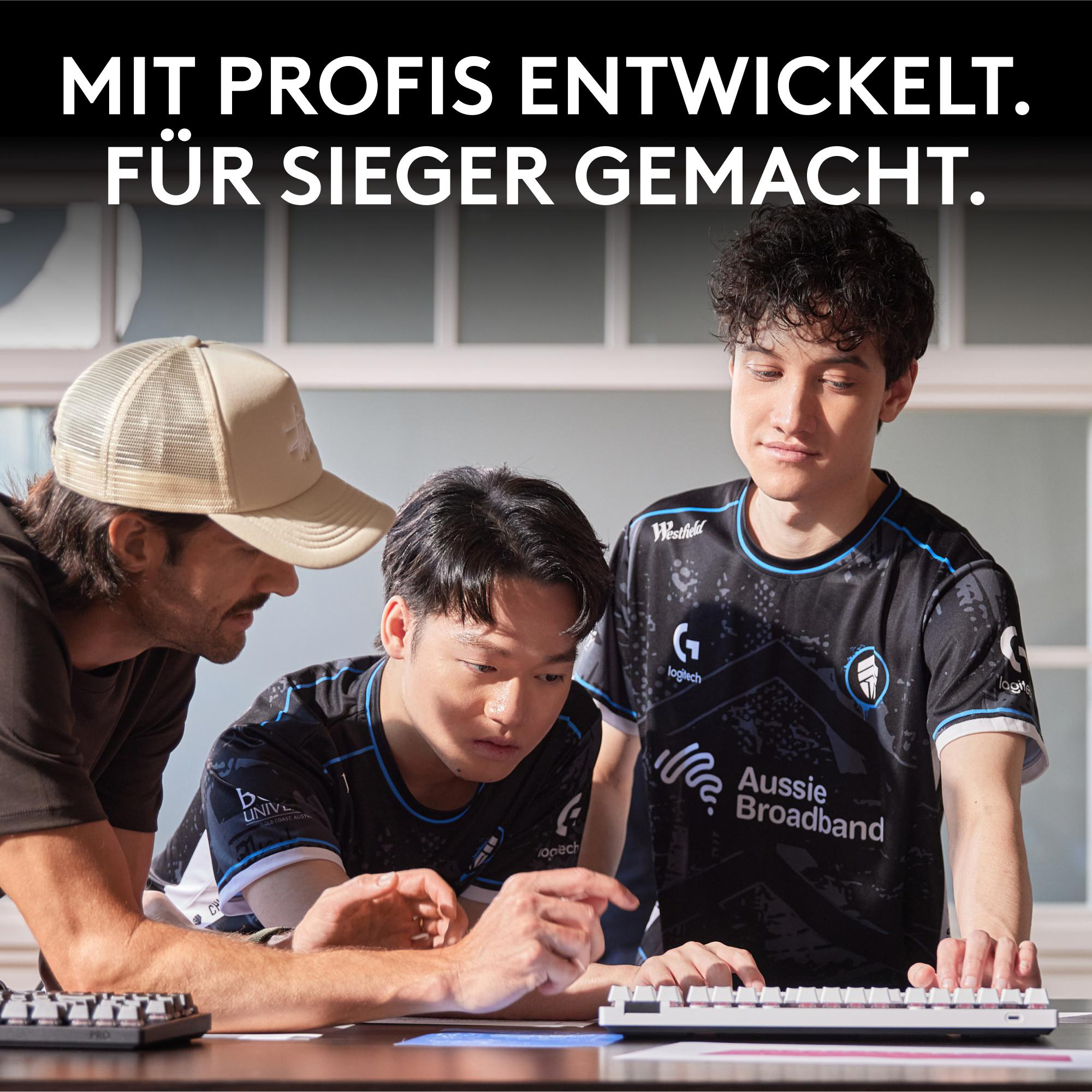 Drei Männer betrachten eine weiße Tastatur. Zwei tragen Trikots mit Logos. Einer trägt eine Kappe.
