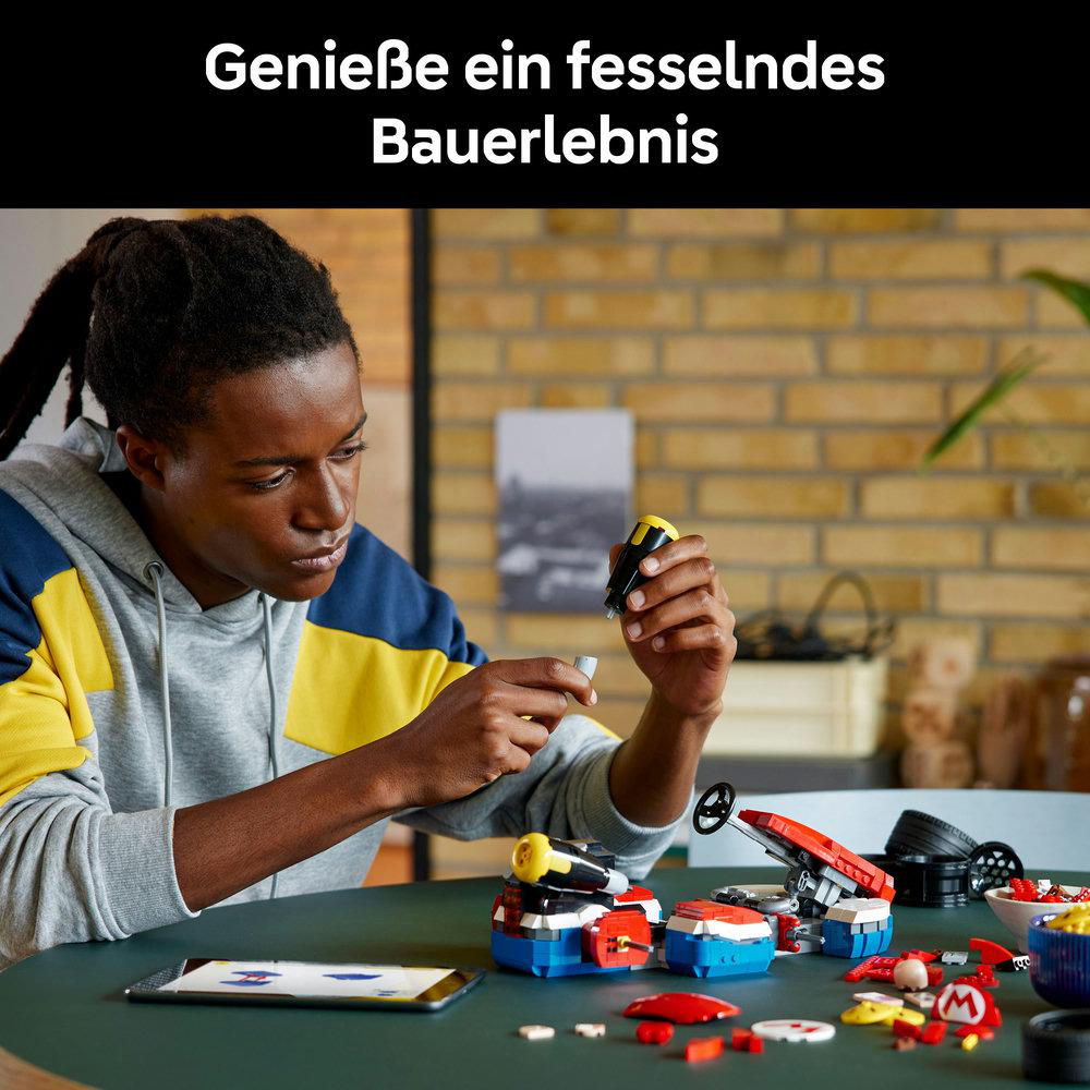 Ein junger Mann baut ein Spielzeugauto aus bunten Blöcken. Er hält ein kleines schwarzes und gelbes Teil. Auf dem Tisch liegen weitere Spielsachen.