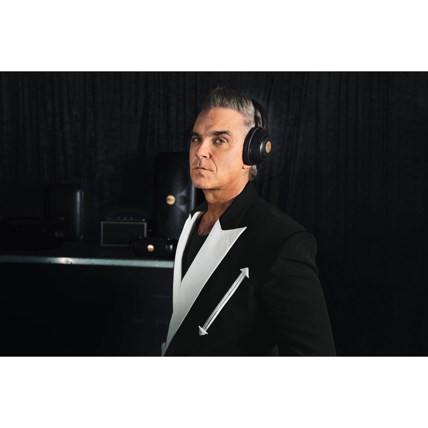 Robbie Williams wearing headphones and a black suit with white lapels. Dark background with studio equipment visible.
