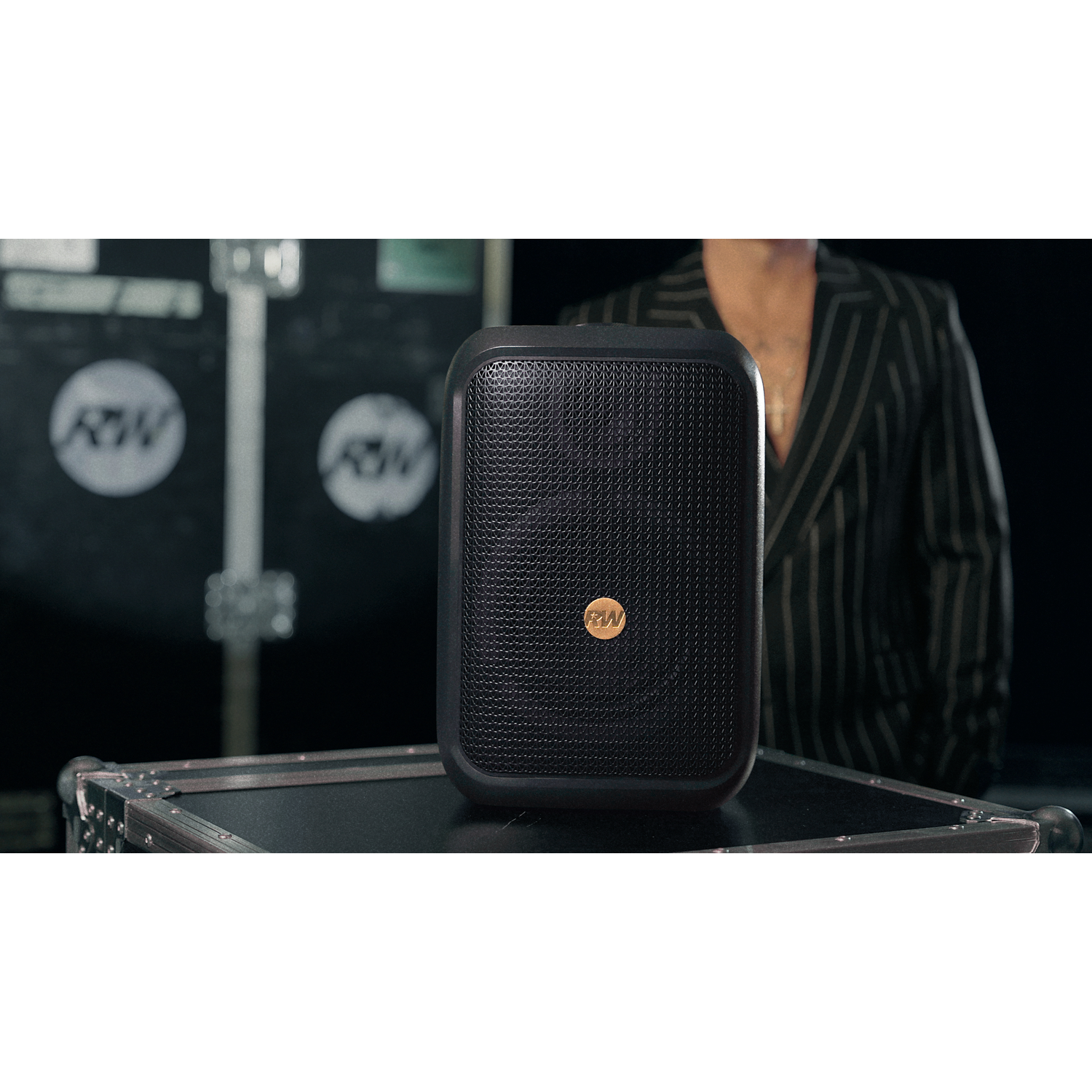 A black speaker with a gold logo sits on a black case. A person in a striped suit stands in the background.