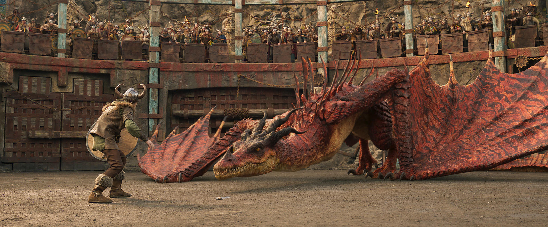 Ein Wikinger steht einem roten Drachen in einer Arena gegenüber. Zuschauer beobachten von oben. Der Drache hat große Flügel, der Wikinger hält einen Schild.