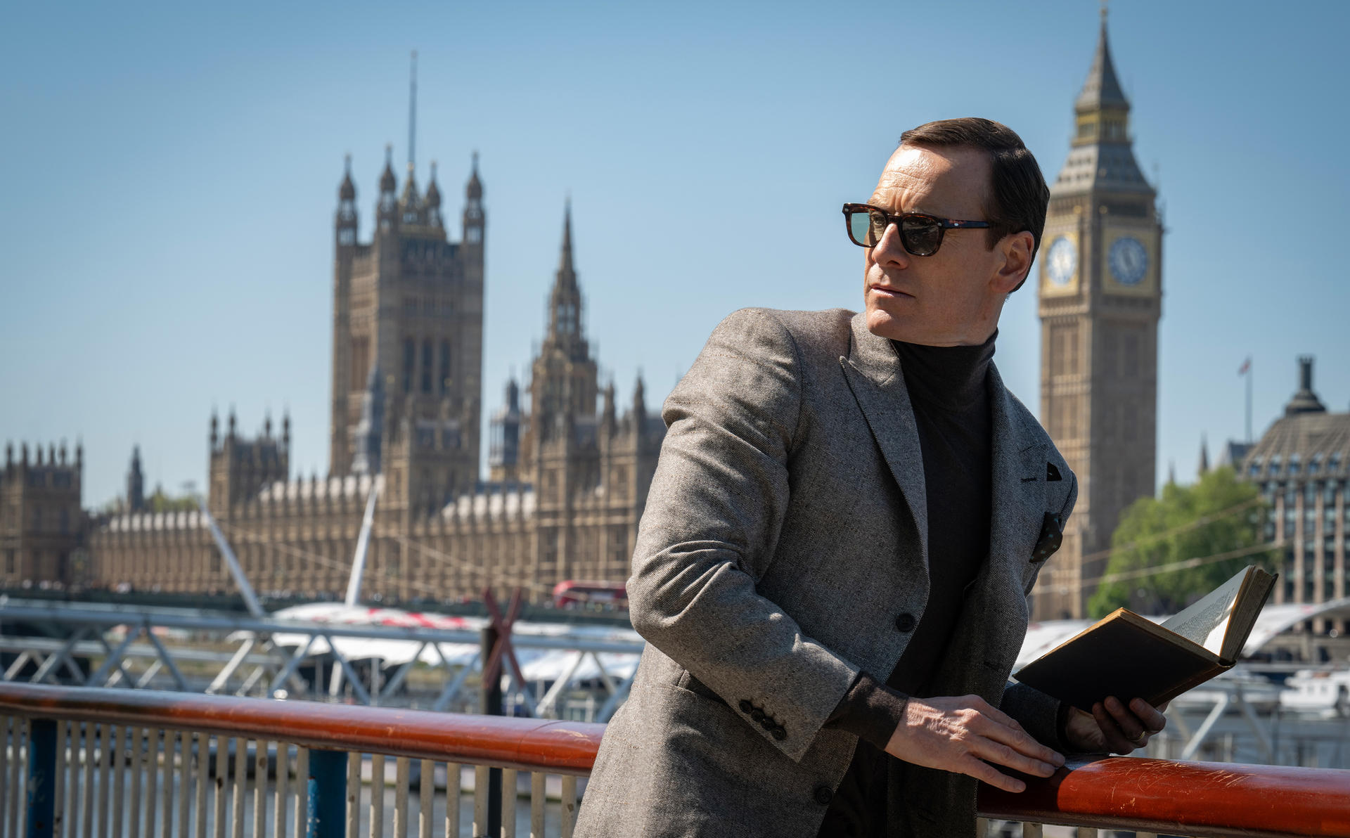 Mann mit Sonnenbrille, grauem Jackett und Buch steht an einem Geländer, im Hintergrund Big Ben und das Parlament.