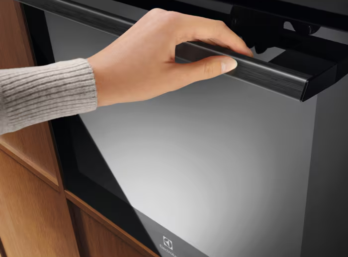 Ręka otwiera drzwi piekarnika. Piekarnik jest czarny, uchwyt ciemny, a otaczająca szafka jest drewniana.