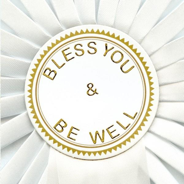 Weiße Rosette mit goldenem Rand und Text: "Bless You & Be Well". Weiße Bänder bilden den Hintergrund.