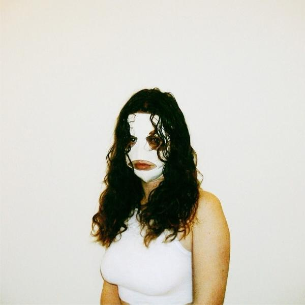 Eine Frau mit lockigem, schwarzem Haar und einer weißen Gesichtsmaske, trägt ein weißes Tanktop vor weißer Wand.