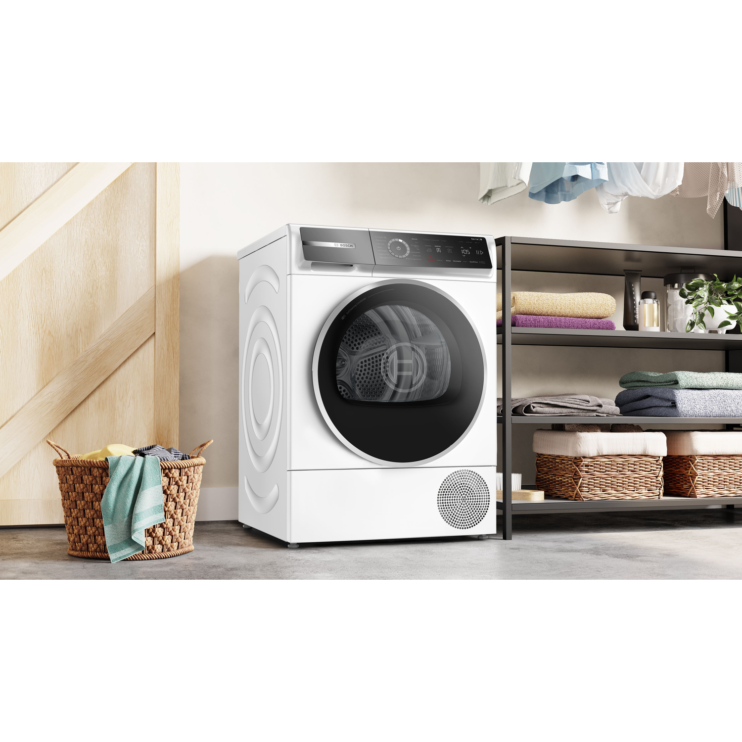 A white dryer is next to a wicker basket with clothes. Shelves with towels are in the background. A wooden door is on the left.