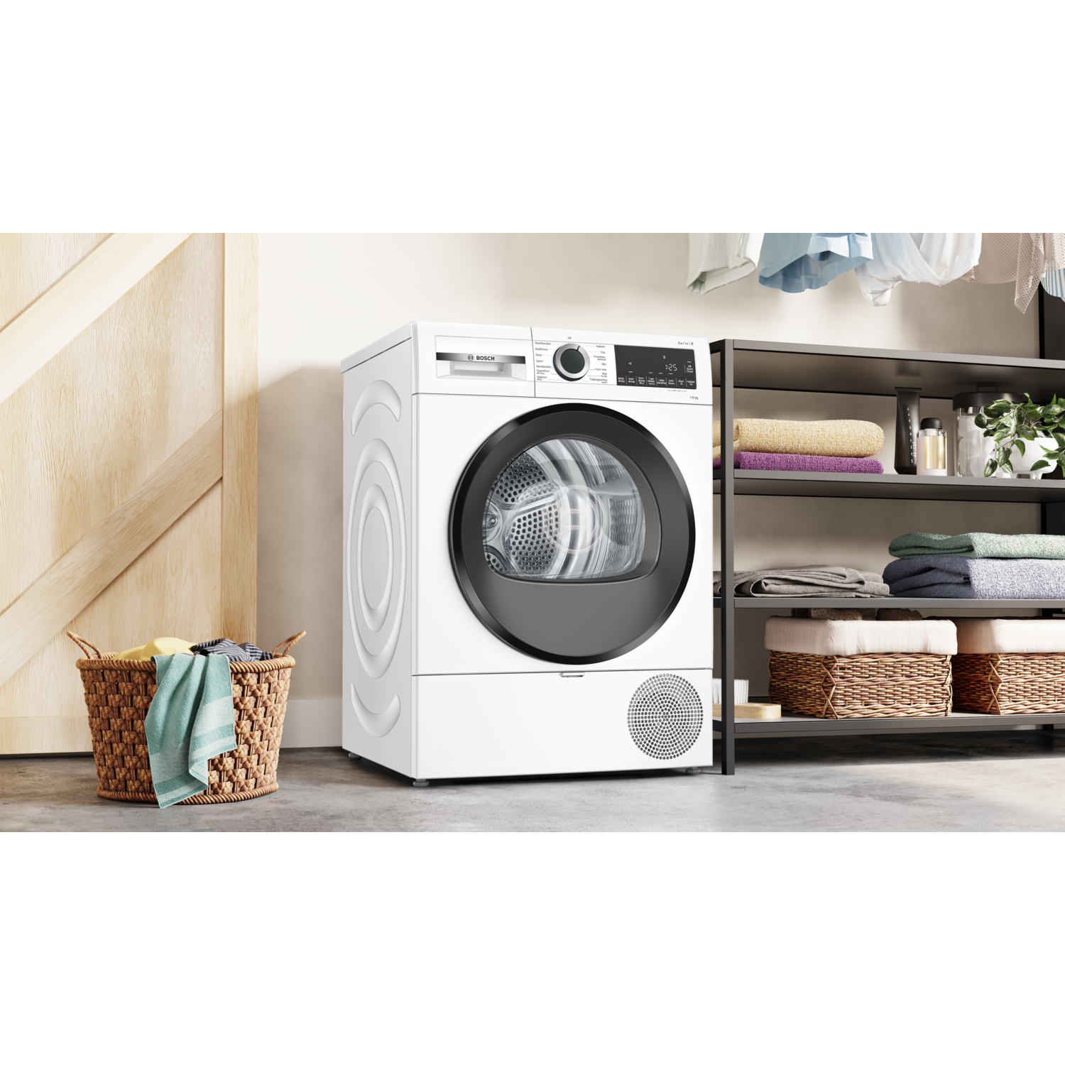 A white dryer stands next to a laundry basket. Shelves hold towels and laundry items. Clothes hang above. A wooden door is on the left.