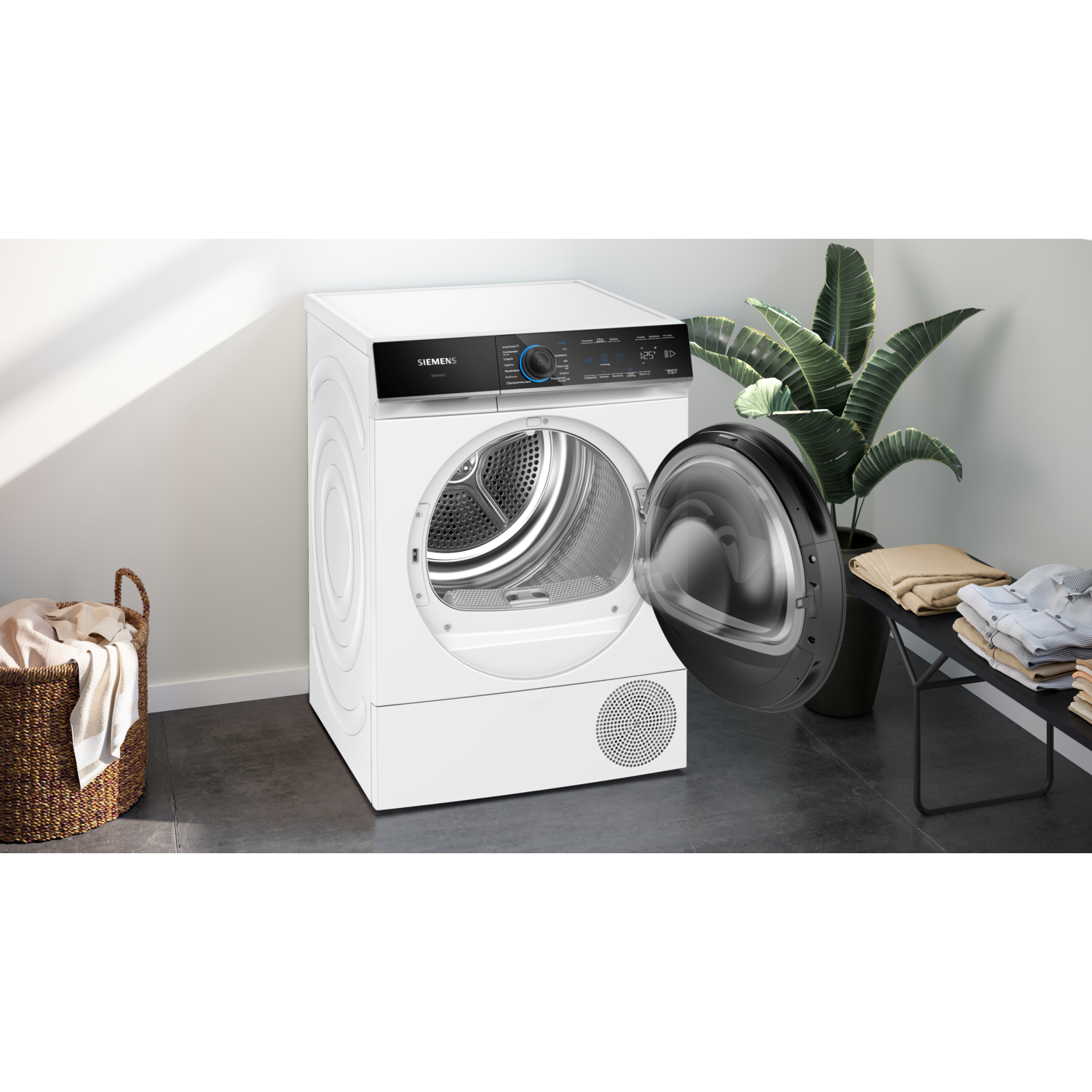 Een witte Siemens droger met een open deur. Een mand met kleding en een plant links en rechts. Een bank met opgevouwen kleding.