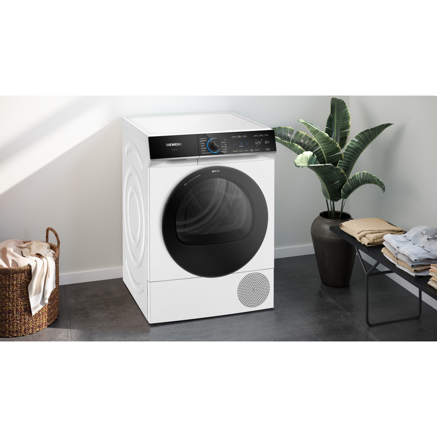 Een witte Siemens droger staat naast een wasmand, een zwarte tafel met opgevouwen kleding en een potplant.