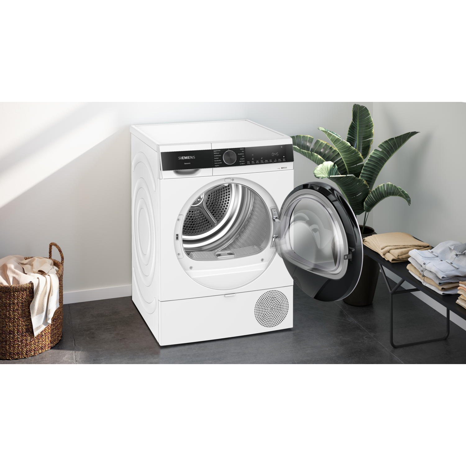 A white Siemens dryer with an open door. A basket of clothes is on the left. A plant and folded clothes are on the right.