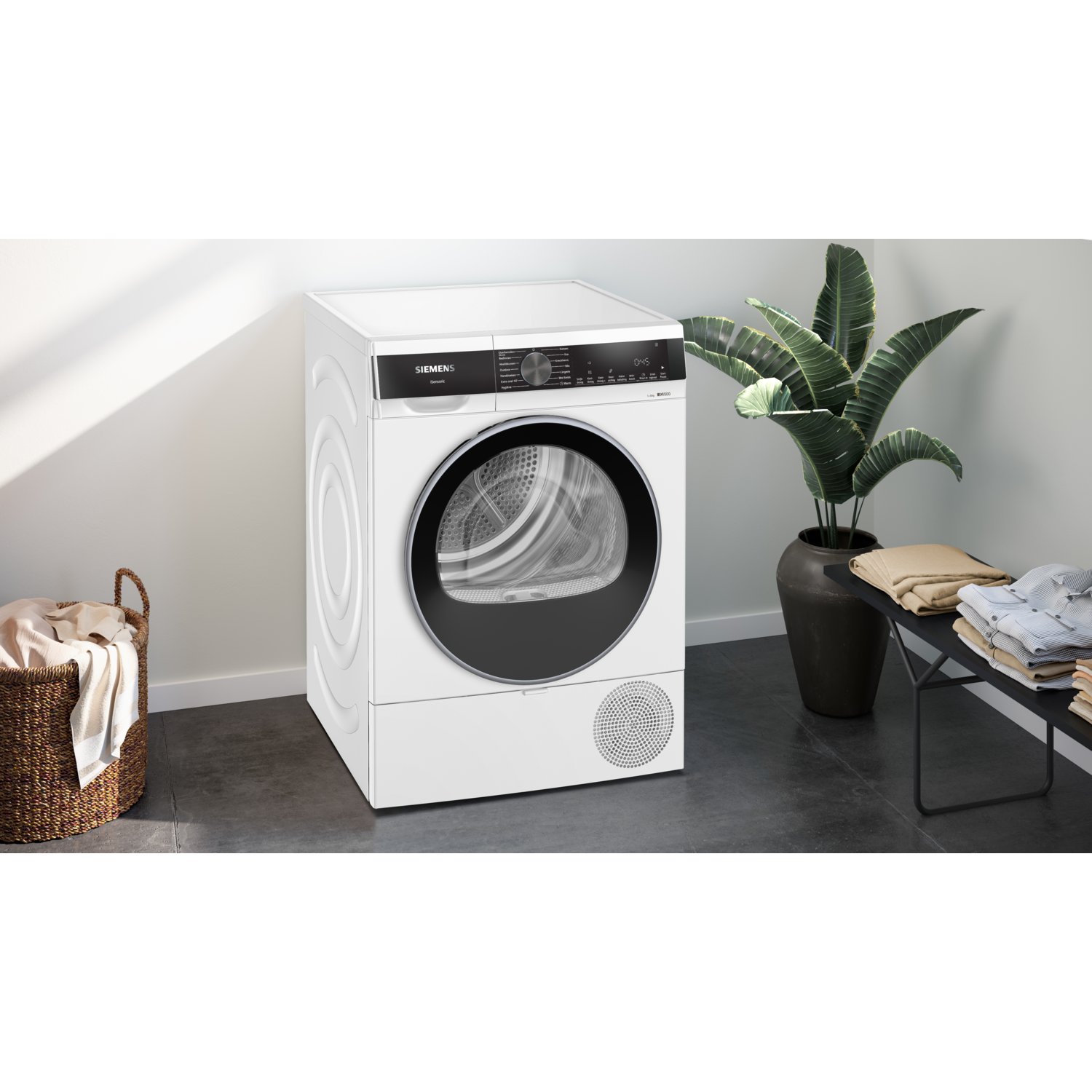 A white Siemens dryer stands in a room with a plant and laundry basket. Clean clothes are on a black bench.