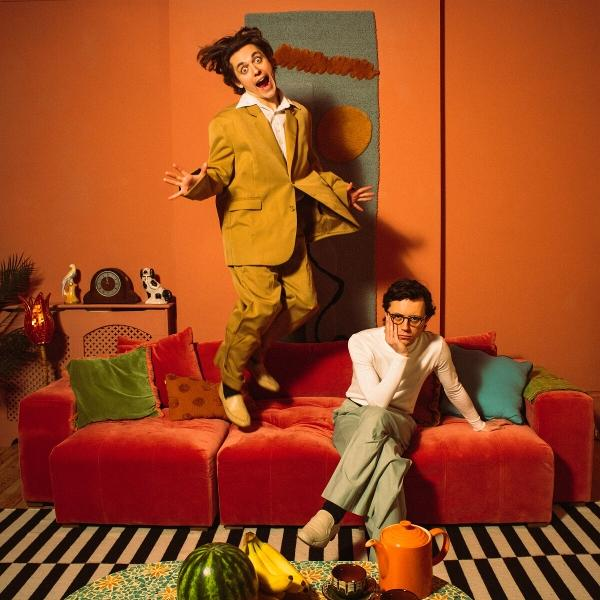 Mann in gelbem Anzug springt über rotes Sofa. Mann mit Brille sitzt auf dem Sofa. Wassermelone, Bananen und Teekanne auf dem Tisch.
