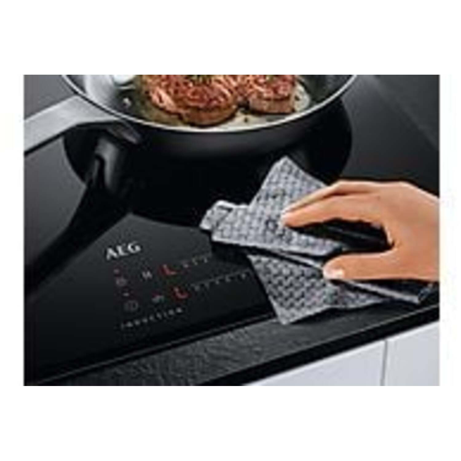 A hand wipes a black induction cooktop with a gray cloth. A pan with cooked meat sits on the cooktop.