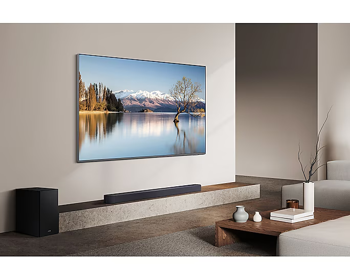 Egy nagyméretű TV-készülék egy tóparti jelenetet mutat hegyekkel. Alatta egy soundbar és egy mélynyomó található. Modern nappali környezet.