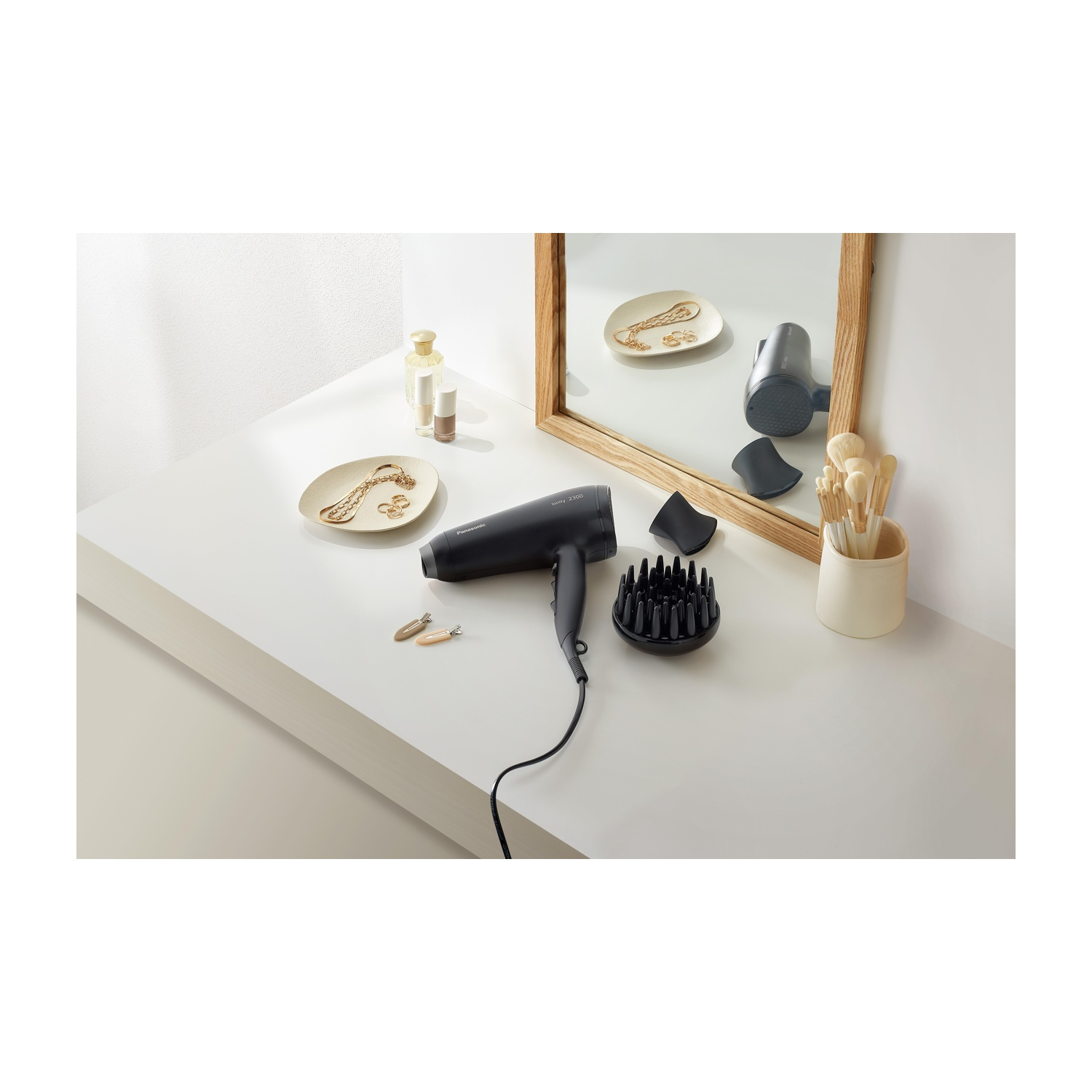 Un asciugacapelli nero, diffusore e accessori su una superficie bianca. Uno specchio riflette gli oggetti. Sono presenti gioielli e trucco.
