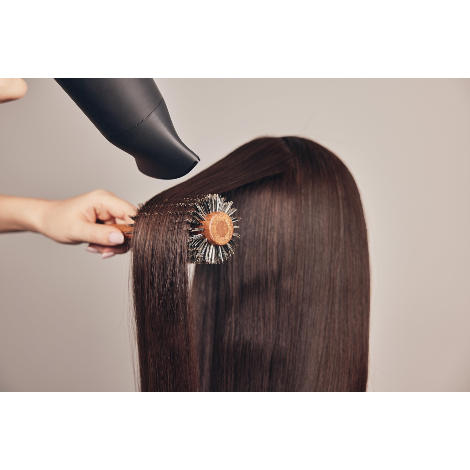 Una persona asciuga con il phon capelli lunghi, lisci e castani con un asciugacapelli nero e una spazzola rotonda.