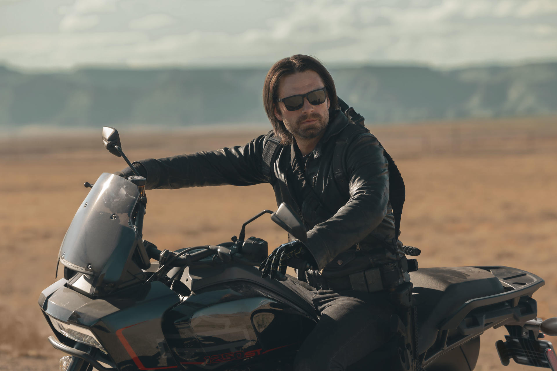 Mann mit Sonnenbrille auf einem Motorrad in einer Wüstenlandschaft. Er trägt eine schwarze Lederjacke und Handschuhe. Das Motorrad ist schwarz.