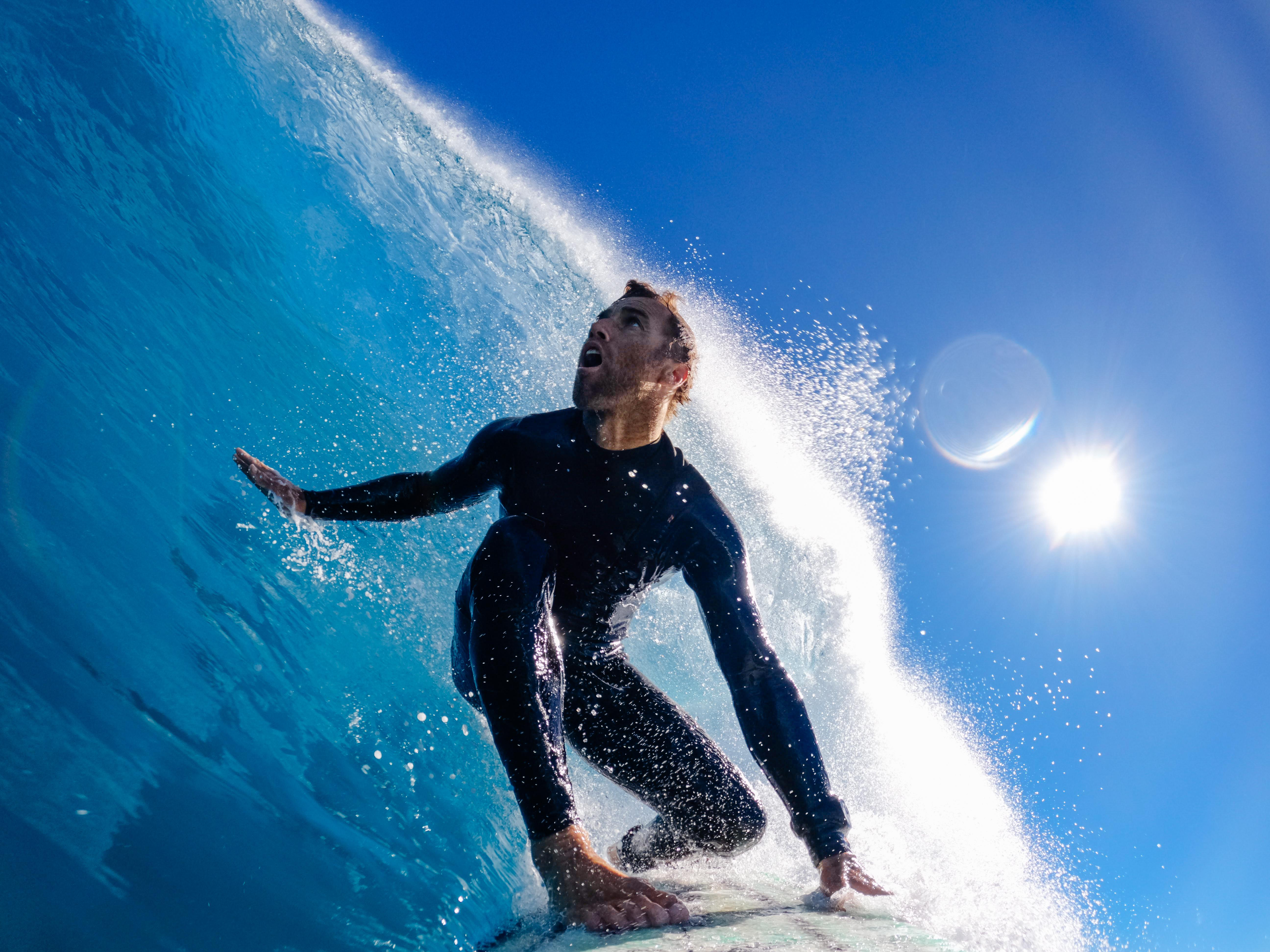 Ein Surfer in einem schwarzen Neoprenanzug reitet eine große Welle unter blauem Himmel. Die Welle bricht über ihm.