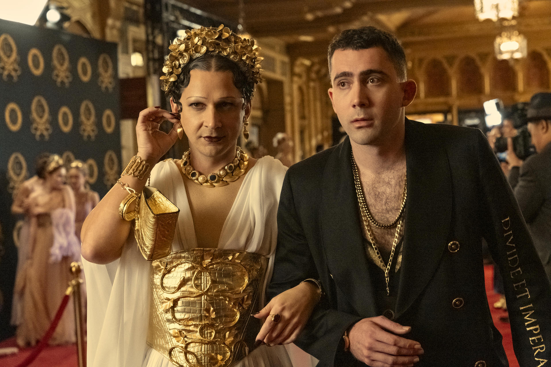 Zwei Personen in formeller Kleidung. Einer trägt goldene Accessoires, der andere einen schwarzen Anzug. Der Hintergrund hat ein Muster und andere Personen.