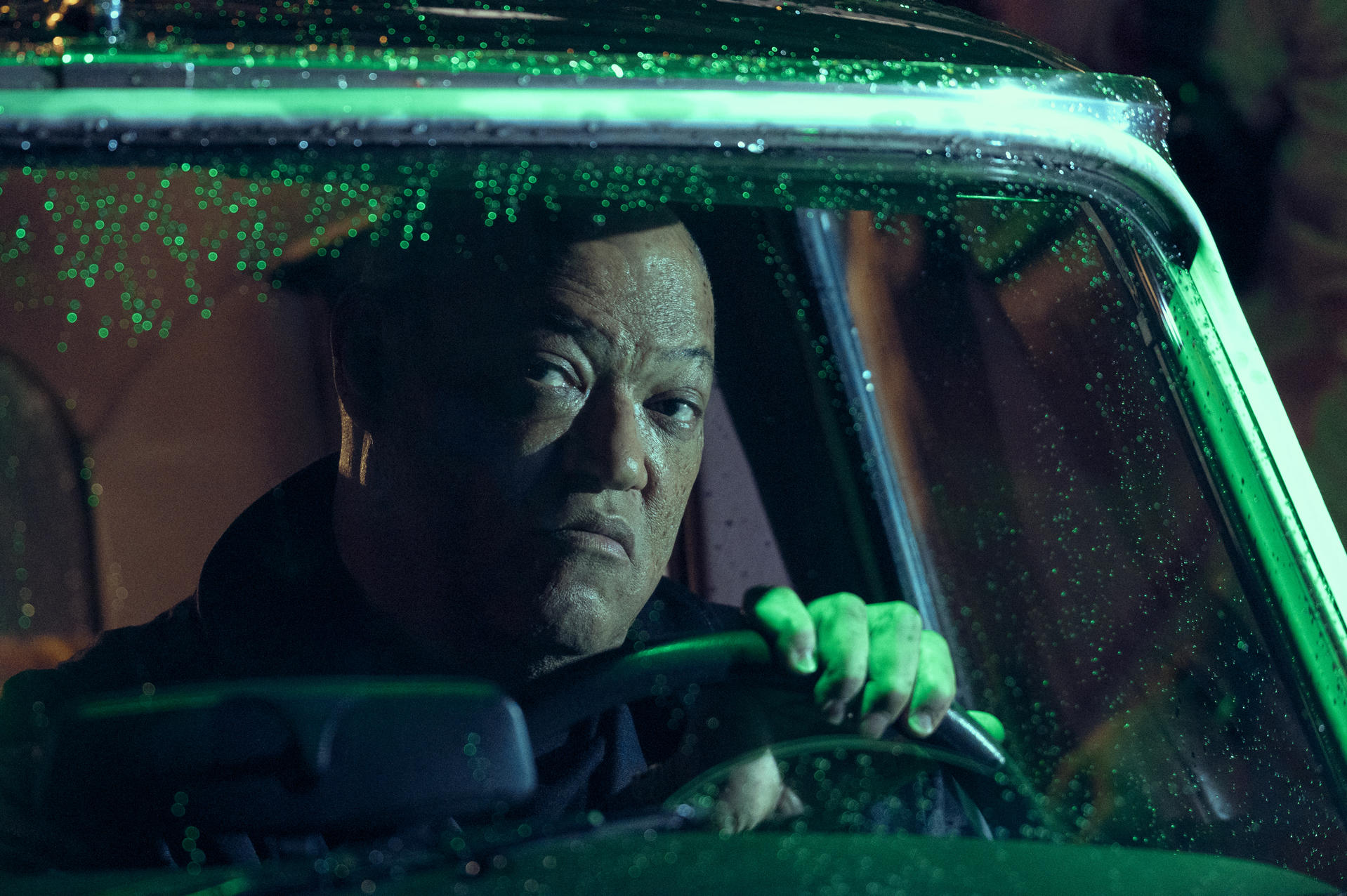 Ein Mann fährt im Regen Auto. Er hält das Lenkrad. Das Auto hat grüne Lichter und der Mann schaut ernst nach vorne.