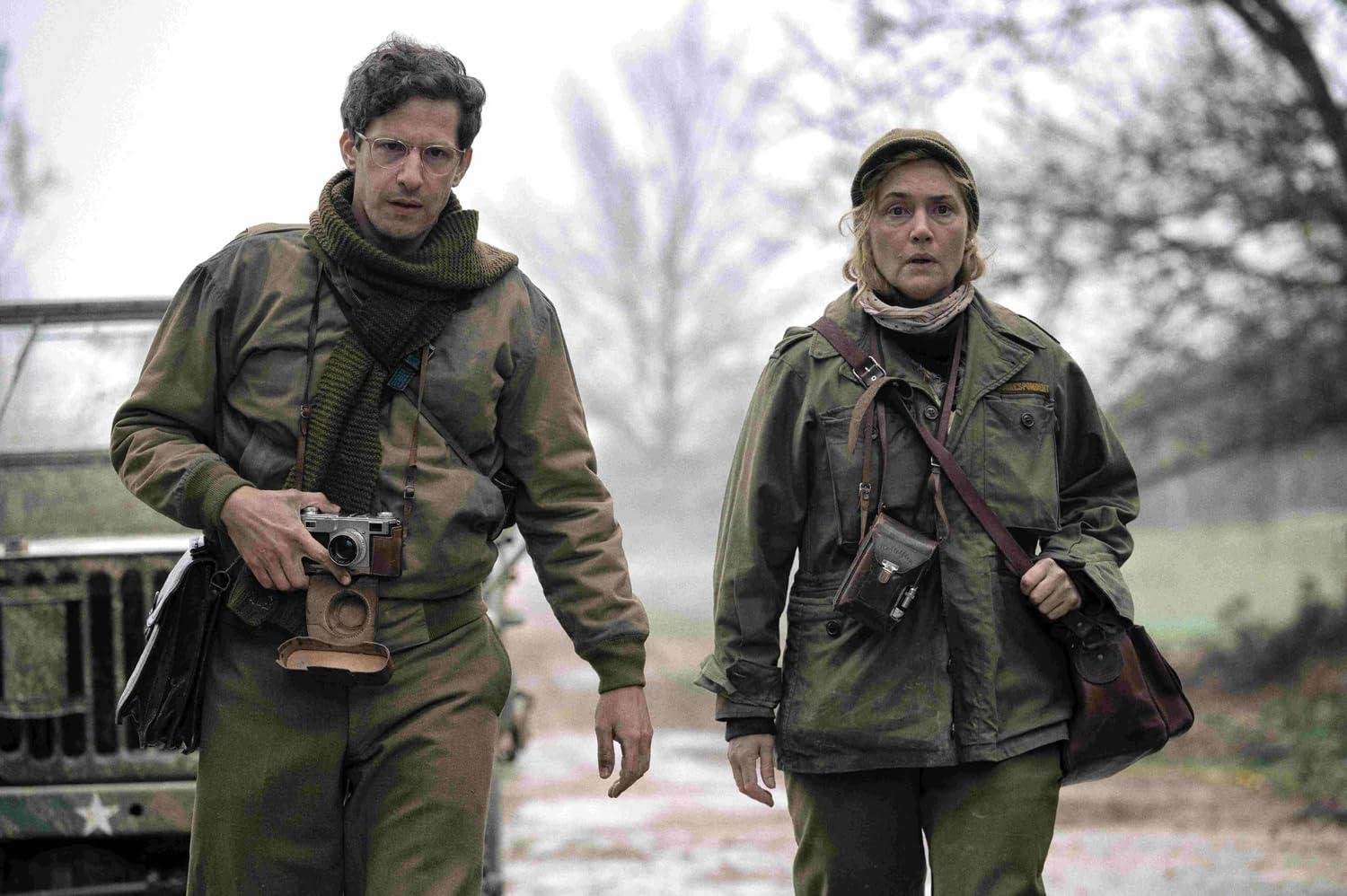Zwei Personen in Militärkleidung gehen vorwärts. Einer hält eine Kamera. Im Hintergrund sind Bäume und ein Jeep zu sehen.