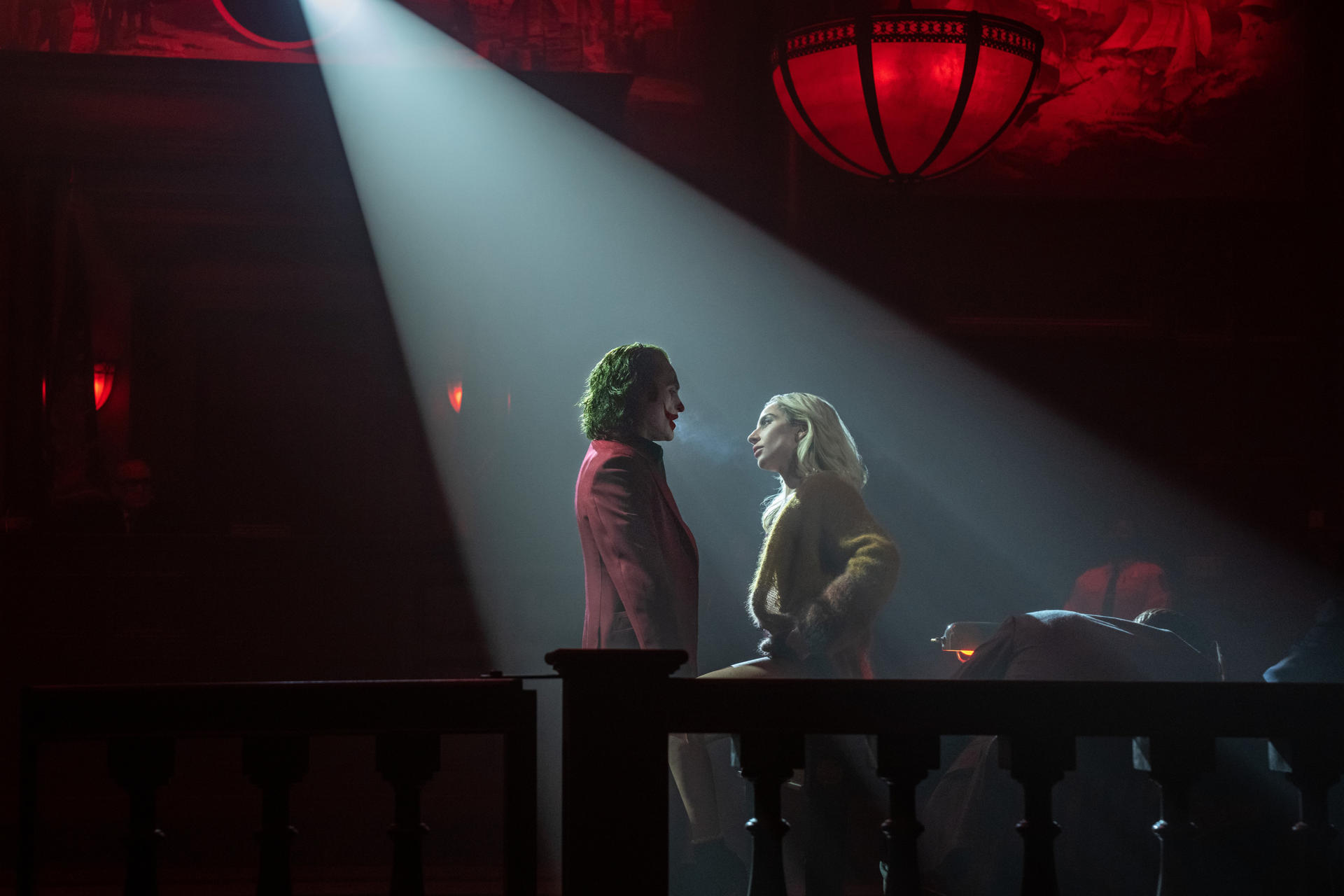 Joker und eine Frau stehen sich unter einem Scheinwerfer gegenüber. Der Joker trägt einen roten Anzug und grünes Haar. Eine rote Lampe hängt oben.