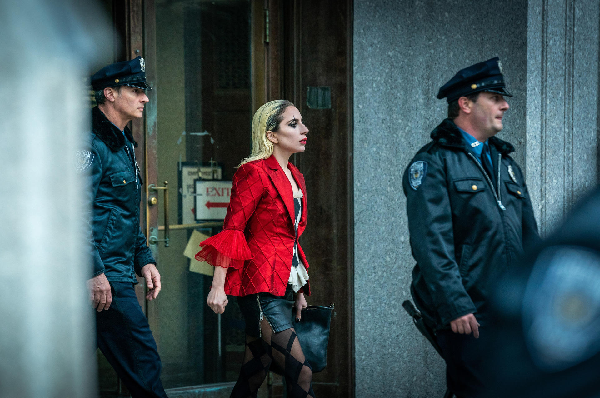 Eine Frau in roter Jacke geht zwischen Polizisten. Sie hat blondes Haar und hält eine schwarze Tasche. Der Hintergrund ist ein Gebäude.