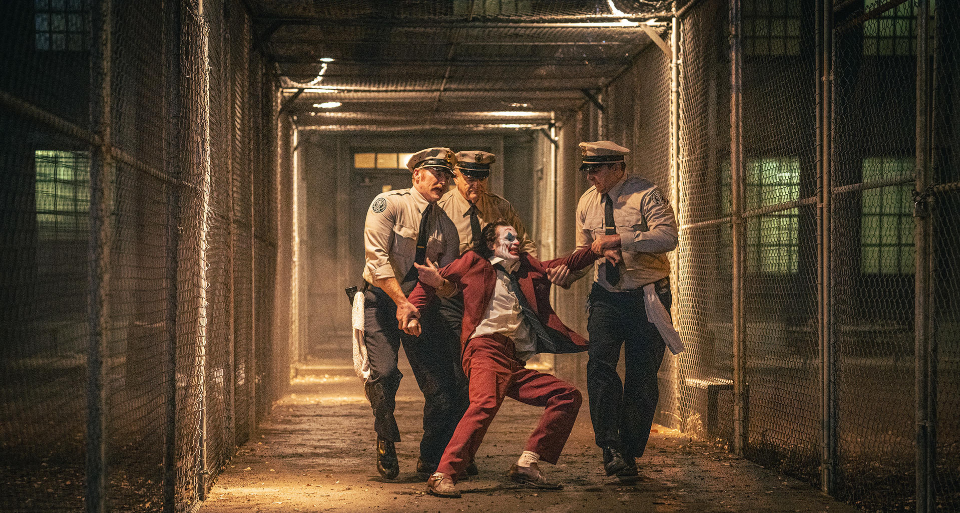 Joker in rotem Anzug wird von drei Polizisten in einem Gefängniskorridor eskortiert. Die Wände sind Maschendrahtzäune.