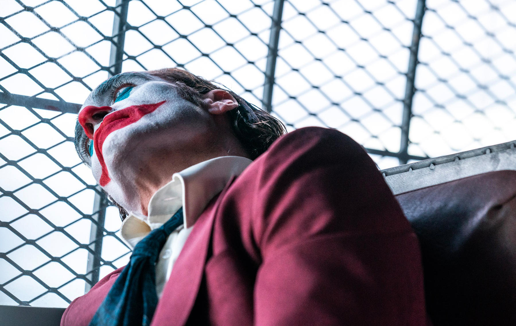 Eine Person mit Joker-Make-up blickt nach oben. Rote Lippen, weißes Gesicht und blauer Lidschatten. Trägt einen roten Blazer und eine dunkelblaue Krawatte.