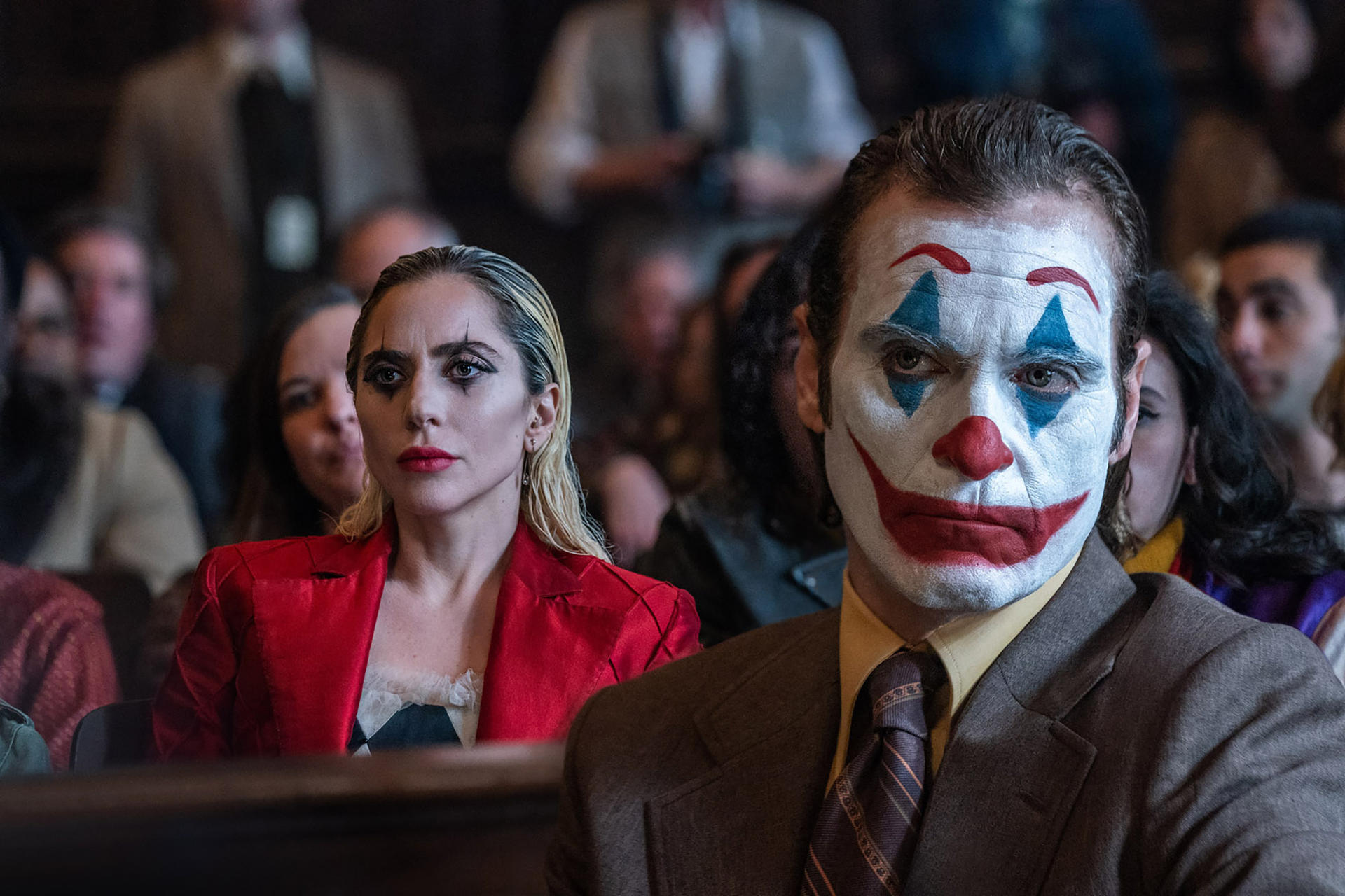 Joker mit Make-up und Lady Gaga in einem roten Blazer sind in einer Menge. Der Joker ist im Vordergrund. Lady Gaga im Hintergrund.