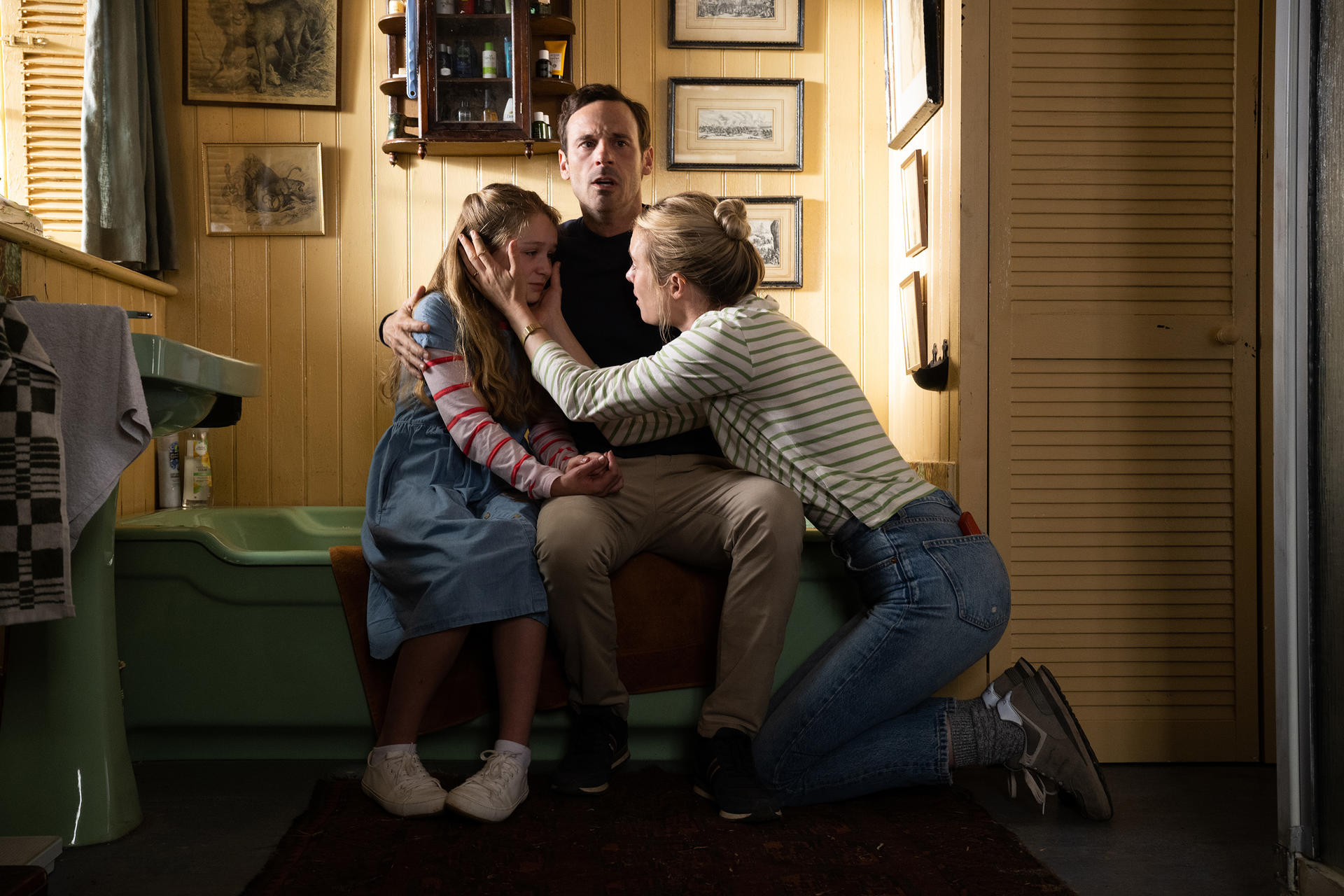 Eine Familie tröstet ein Mädchen in einem Badezimmer. Das Mädchen trägt ein blaues Kleid. Die Frau trägt Jeans und ein gestreiftes Hemd.