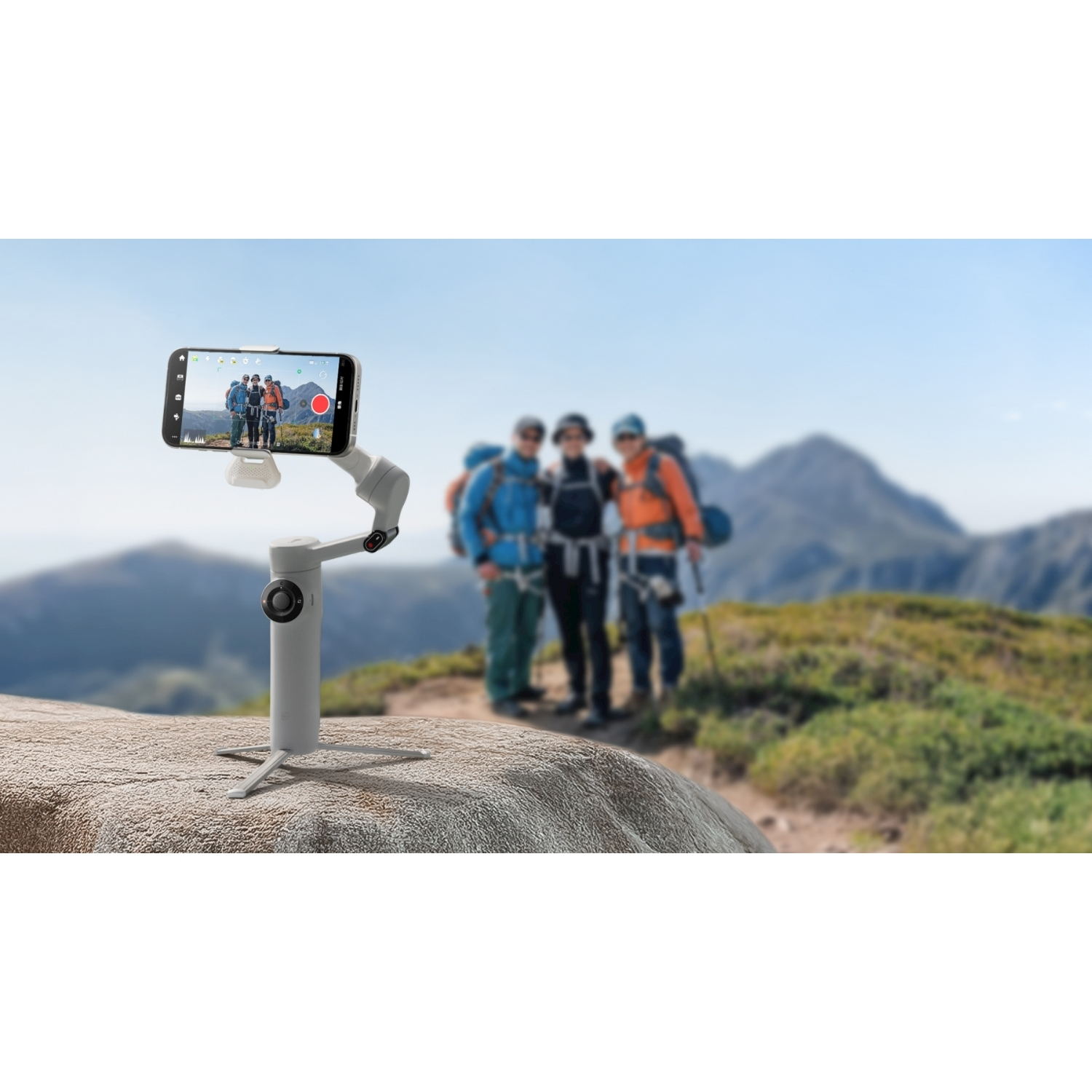 Un telefono su un gimbal riprende tre escursionisti su un sentiero di montagna, con montagne e cielo blu sullo sfondo.