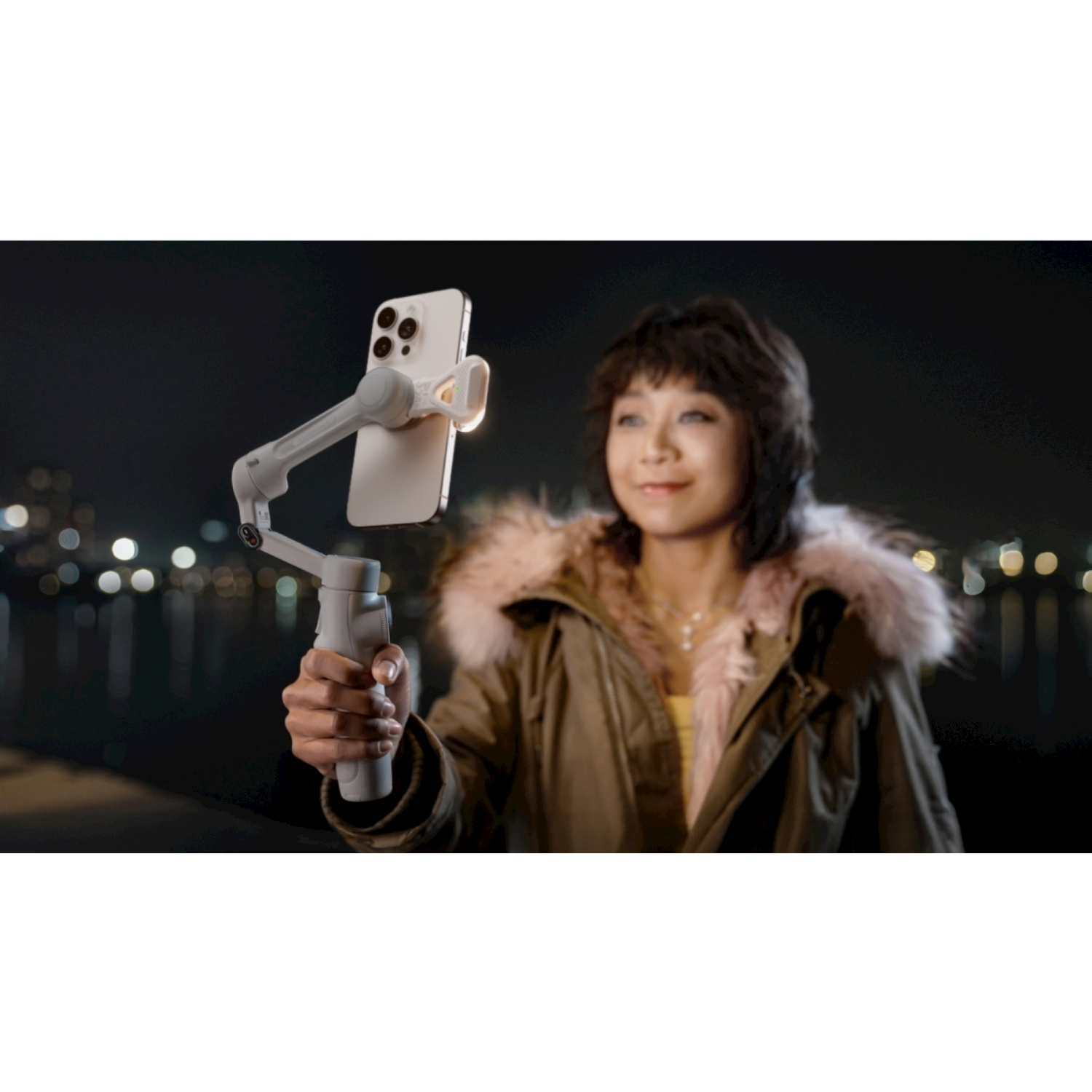 Una donna tiene in mano un telefono su un gimbal. Indossa un cappotto con pelliccia rosa. Scena notturna con luci della città sullo sfondo.