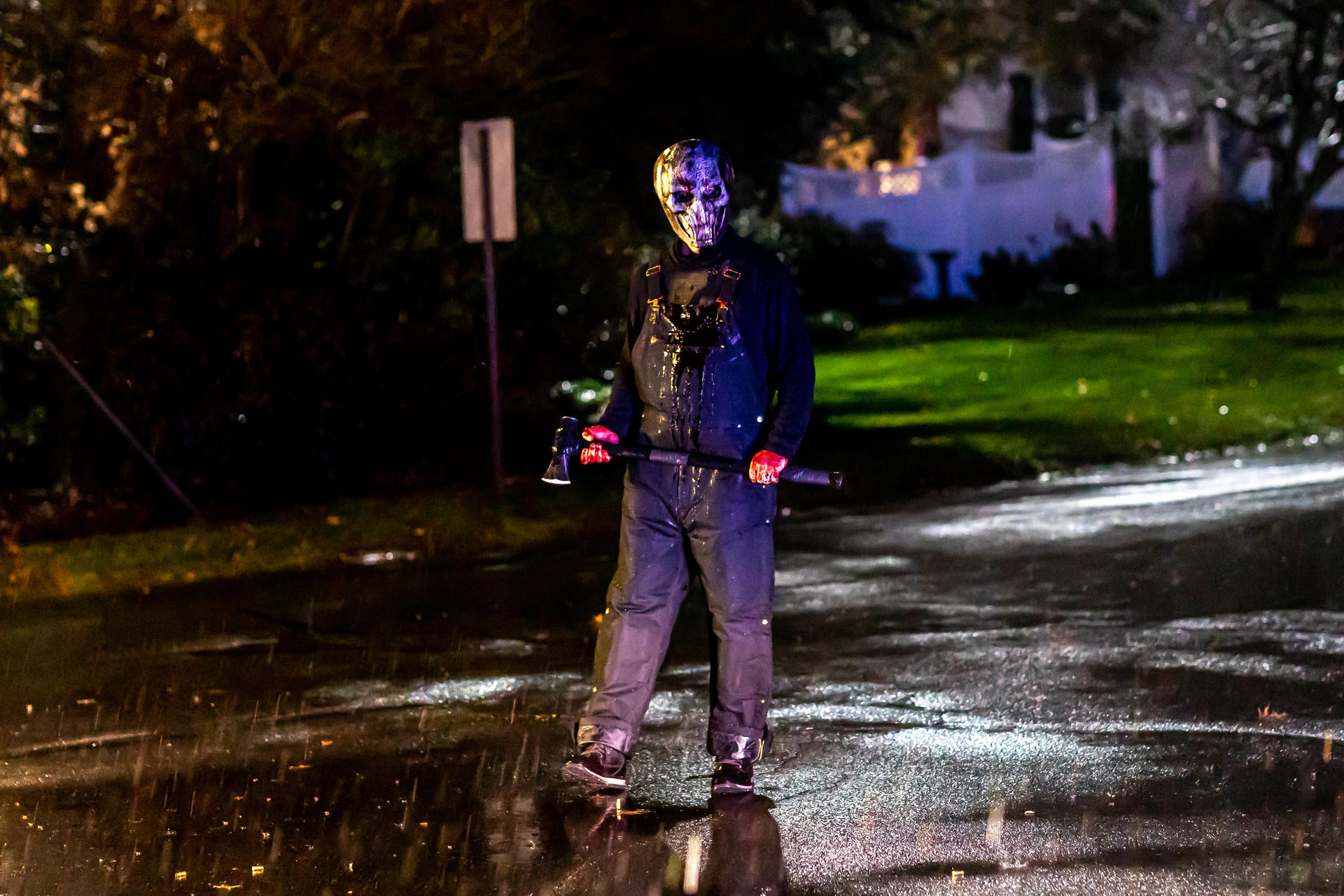 Eine Person mit Maske und Latzhose hält eine Axt. Die Person steht nachts auf einer nassen Straße mit Bäumen und einem Haus im Hintergrund.