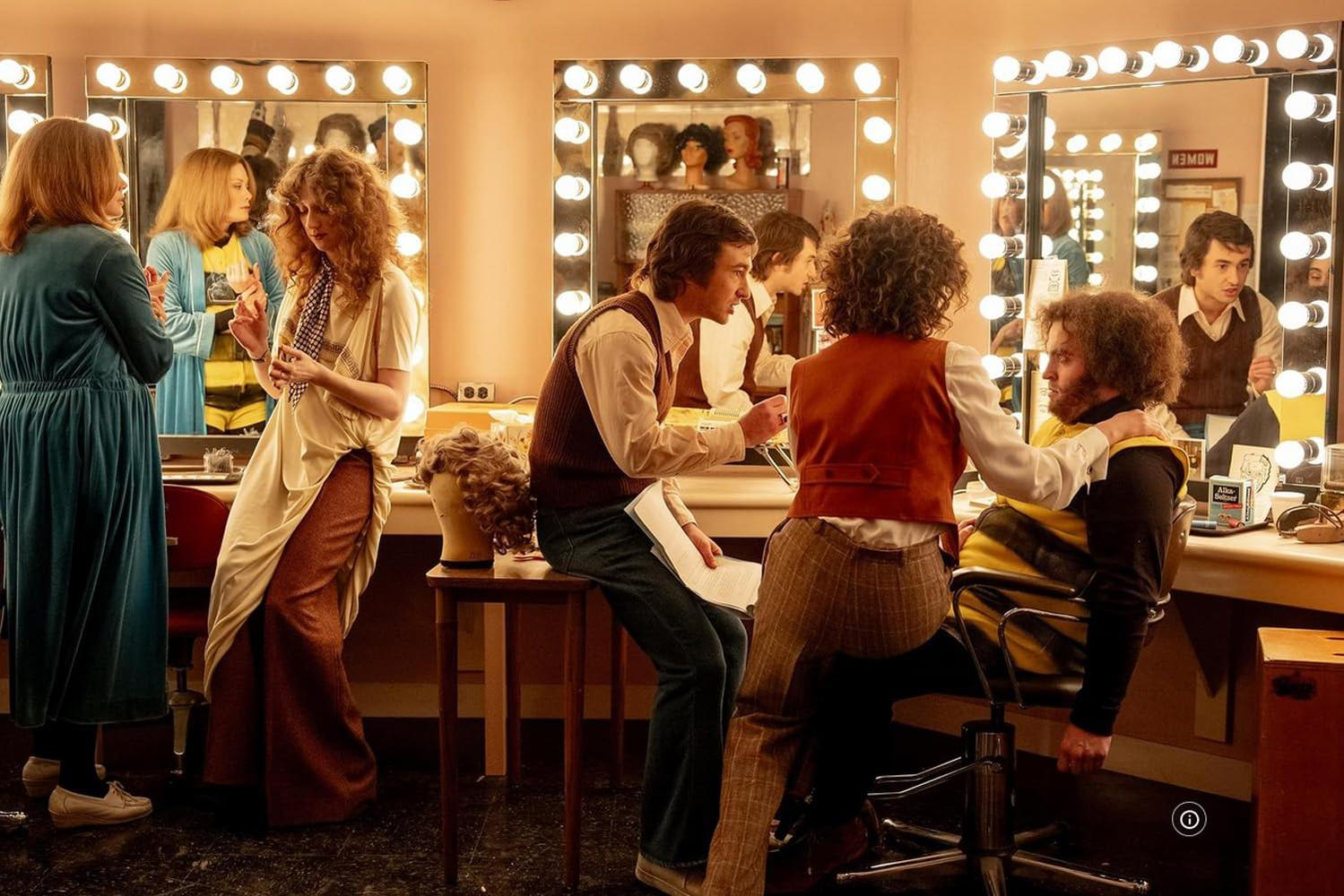 Schauspieler in einer Garderobe. Spiegel mit Lichtern, Perücken und Vintage-Kleidung. Menschen interagieren und bereiten sich auf eine Show vor.