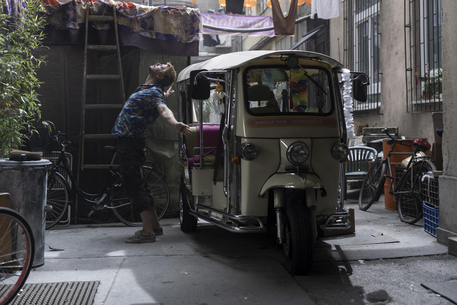 Ein Mann steht in einer Gasse neben einem Tuk-Tuk. Fahrräder lehnen an Wänden. Gebäude und Wäsche hängen im Hintergrund.