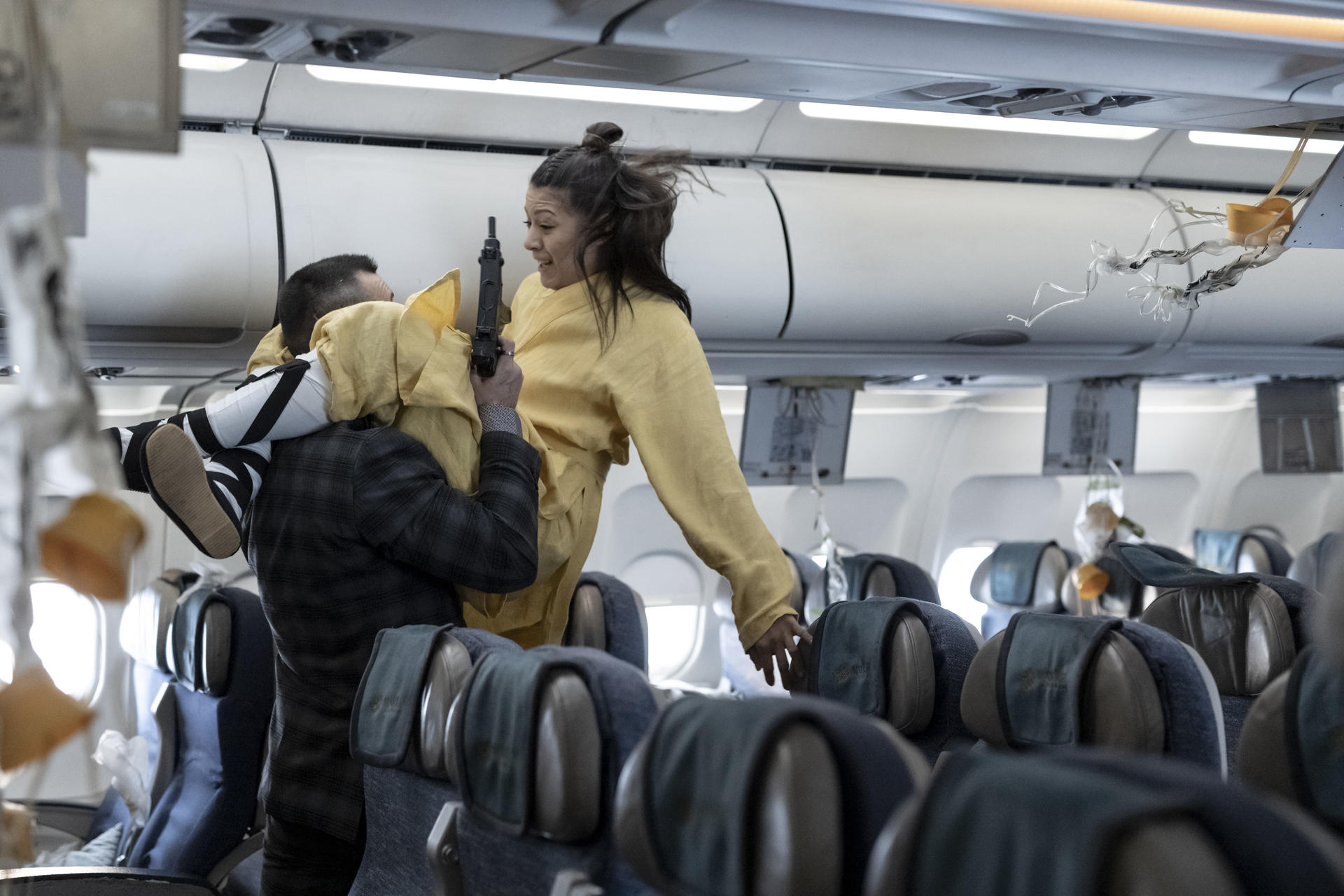 Mann trägt Frau, die eine Waffe hält, durch Flugzeugkabine mit beschädigten Sauerstoffmasken.