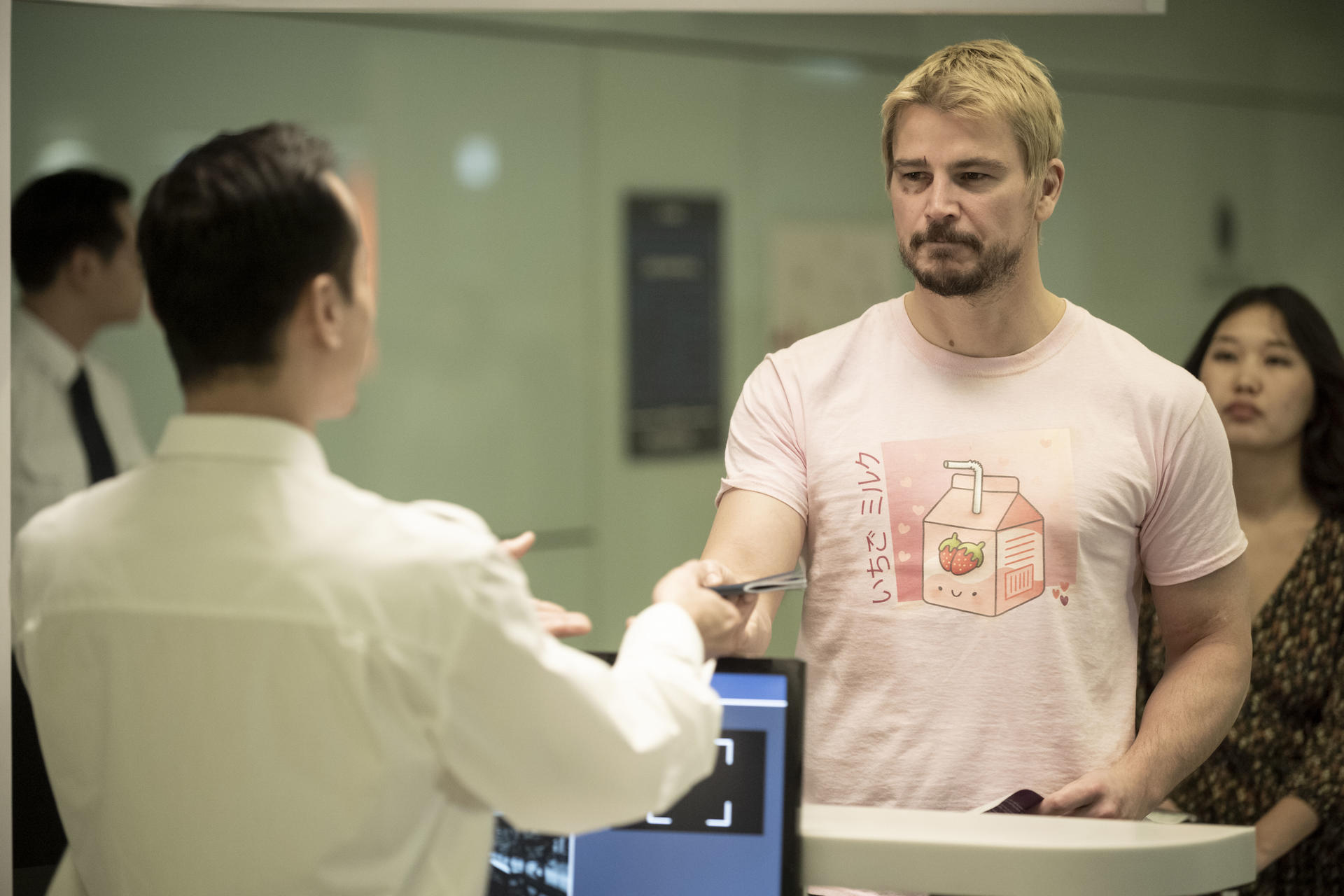Mann in rosa T-Shirt gibt Dokumente an Angestellten hinter einem Schreibtisch.