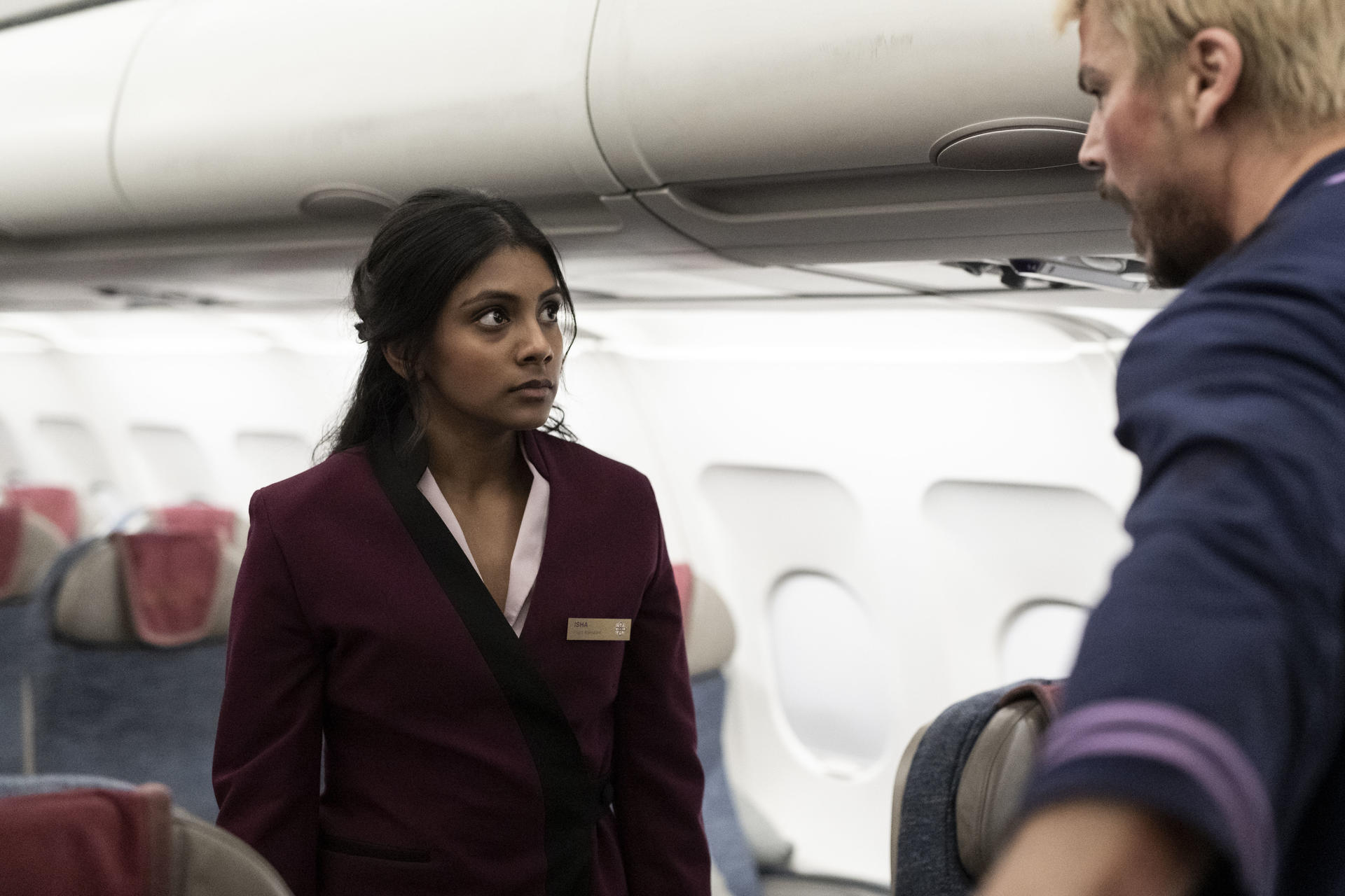Eine Stewardess in bordeauxroter Uniform spricht mit einem Mann im Flugzeug.