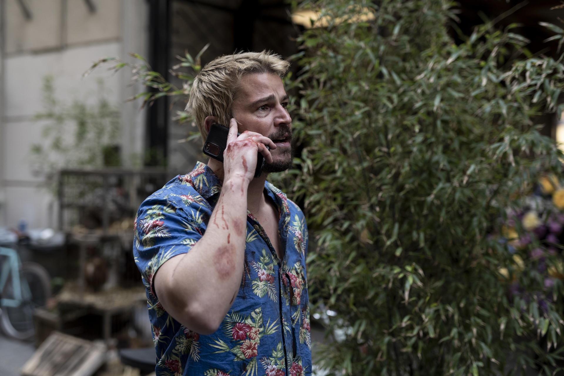 Ein Mann mit blonden Haaren telefoniert, sein Arm ist verletzt und blutig.