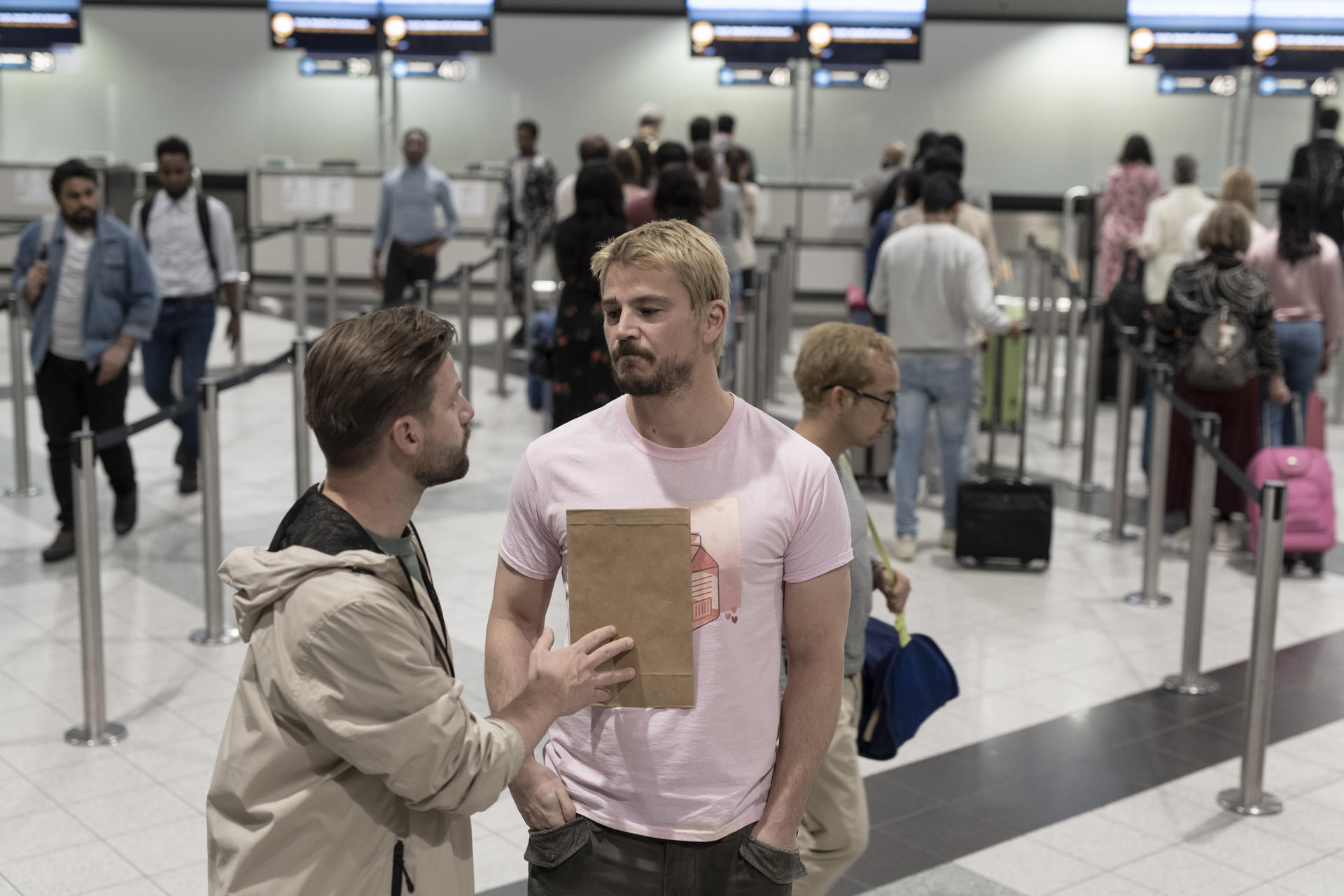 Zwei Männer stehen im Vordergrund, im Hintergrund eine Flughafen-Schlange.